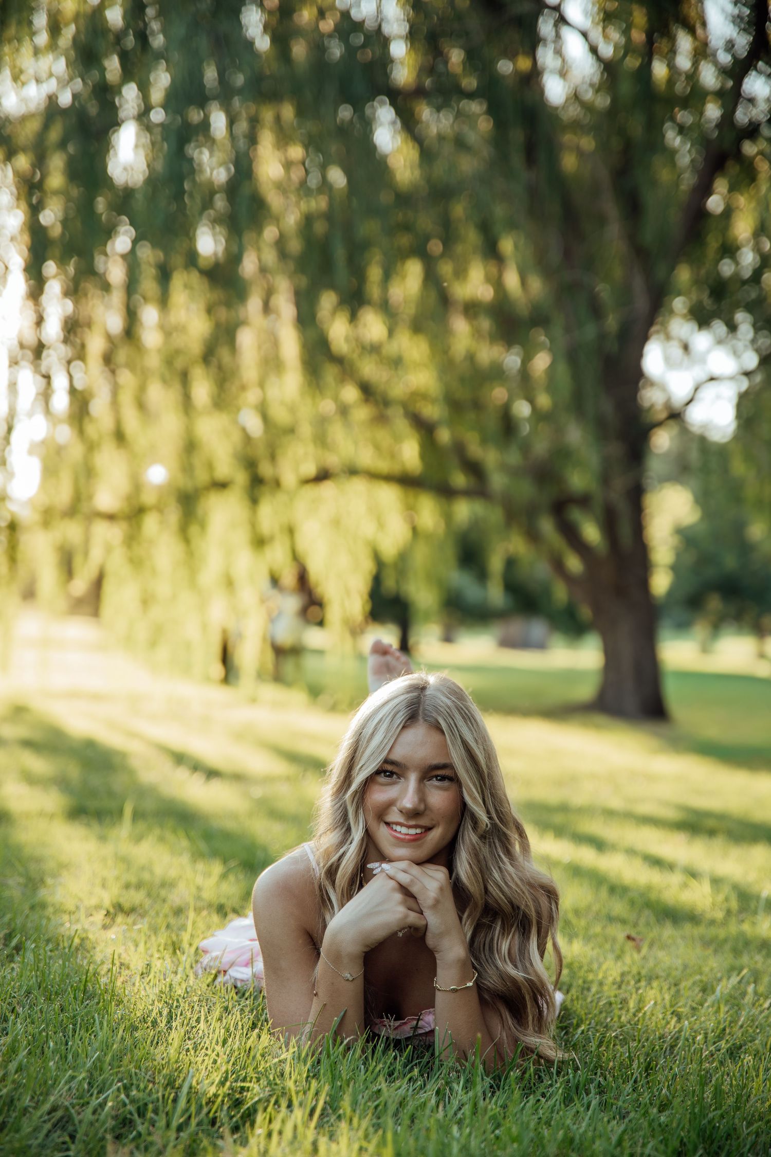 Smiling blonde woman lying on green grass under a weeping willow tree in golden sunlight.