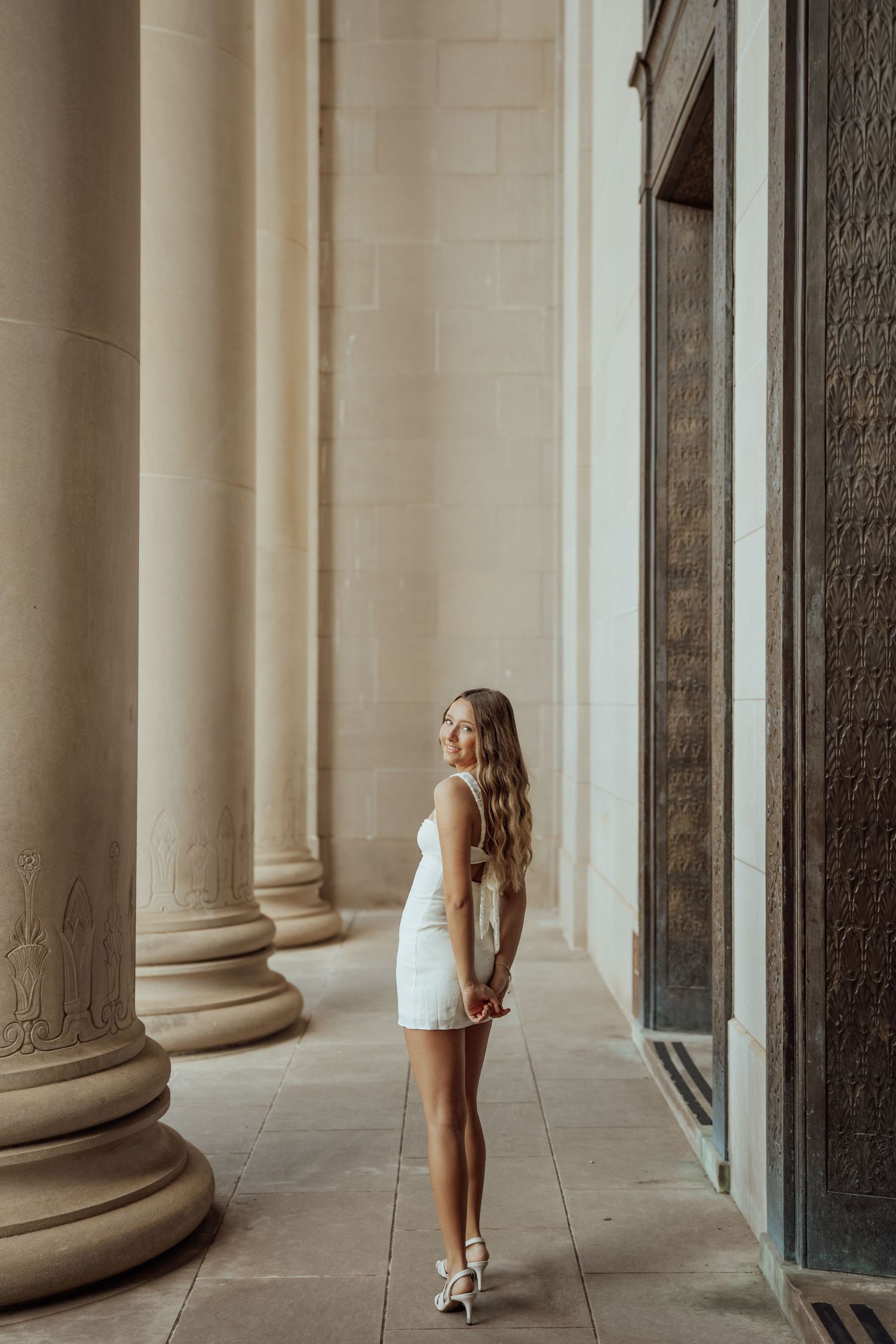 Blonde woman in white mini dress standing among tall stone columns of a classical building.