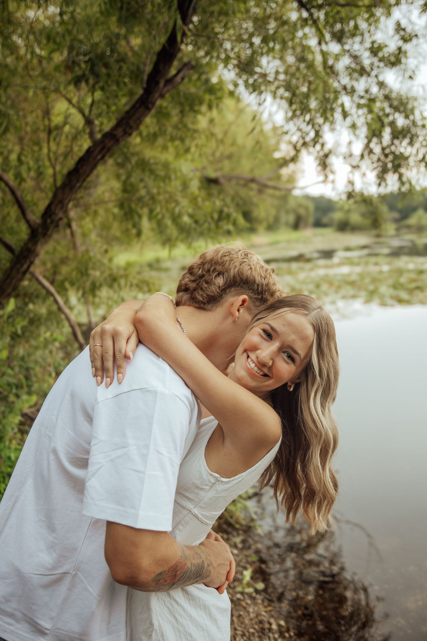 A smiling woman embraces a man near a tranquil pond with green trees in the background.