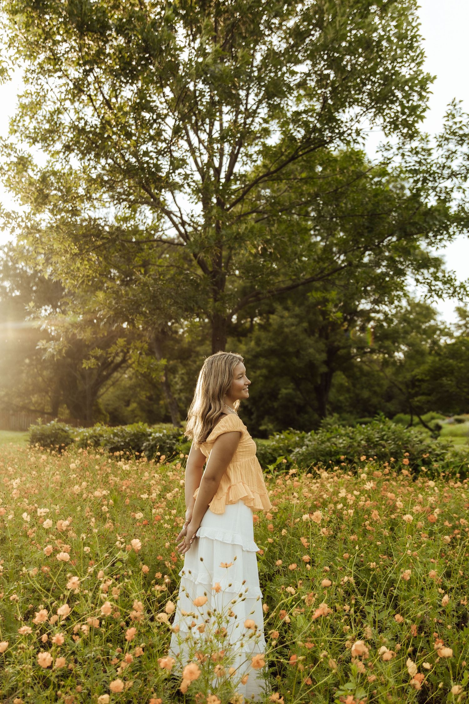 Woman in yellow top and floral skirt standing in a sunlit garden field with blooming flowers and lush green trees.