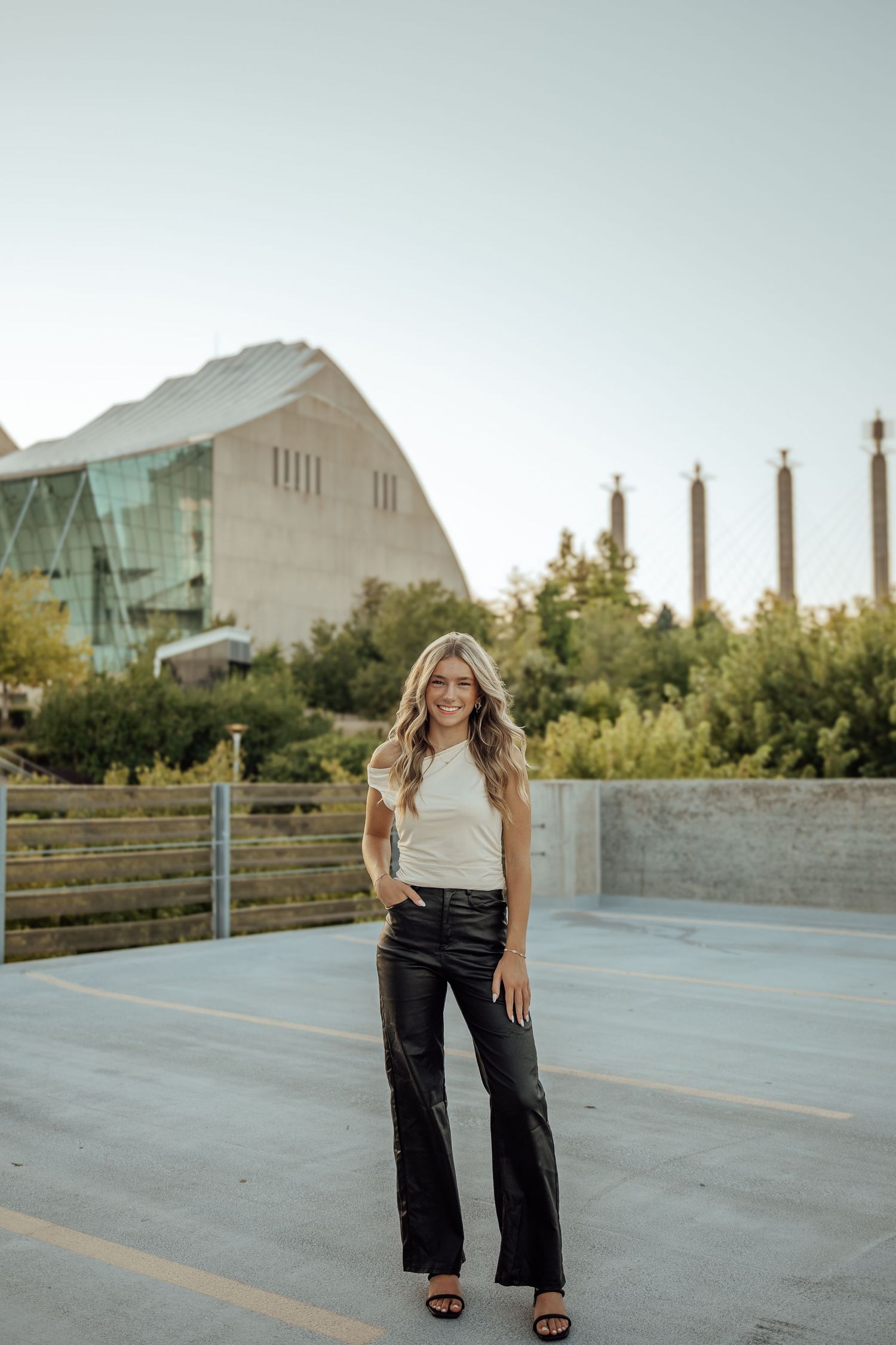 Blonde woman in white top and black leather wide-leg pants smiling on rooftop with modern building behind.