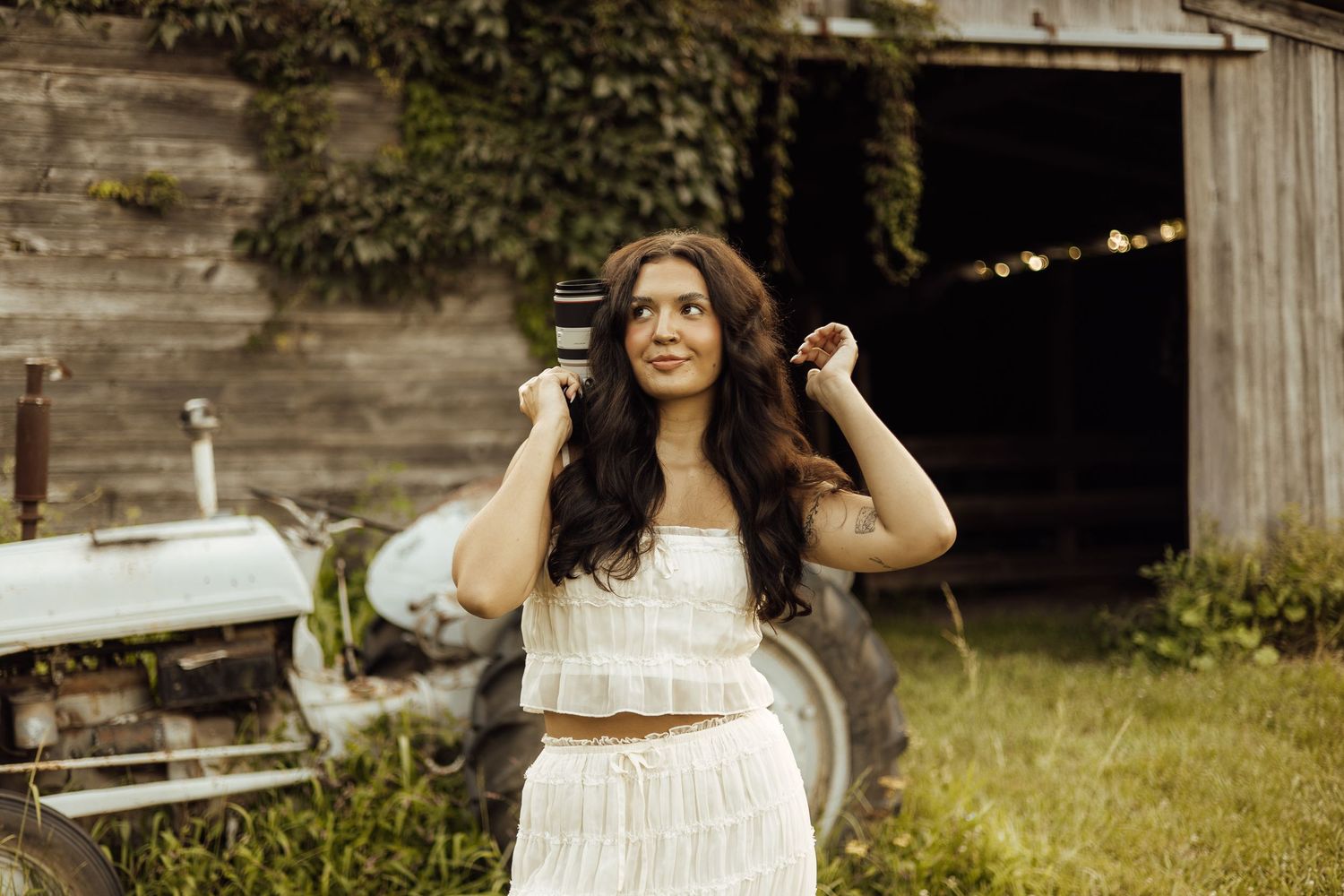 Young woman in white crop top and skirt poses outdoors near rustic barn with vintage tractor and lush greenery.