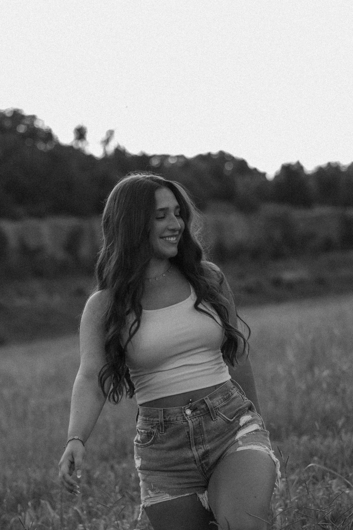 Black and white photo of a young woman with long wavy hair in a crop top and denim shorts smiling in a field.