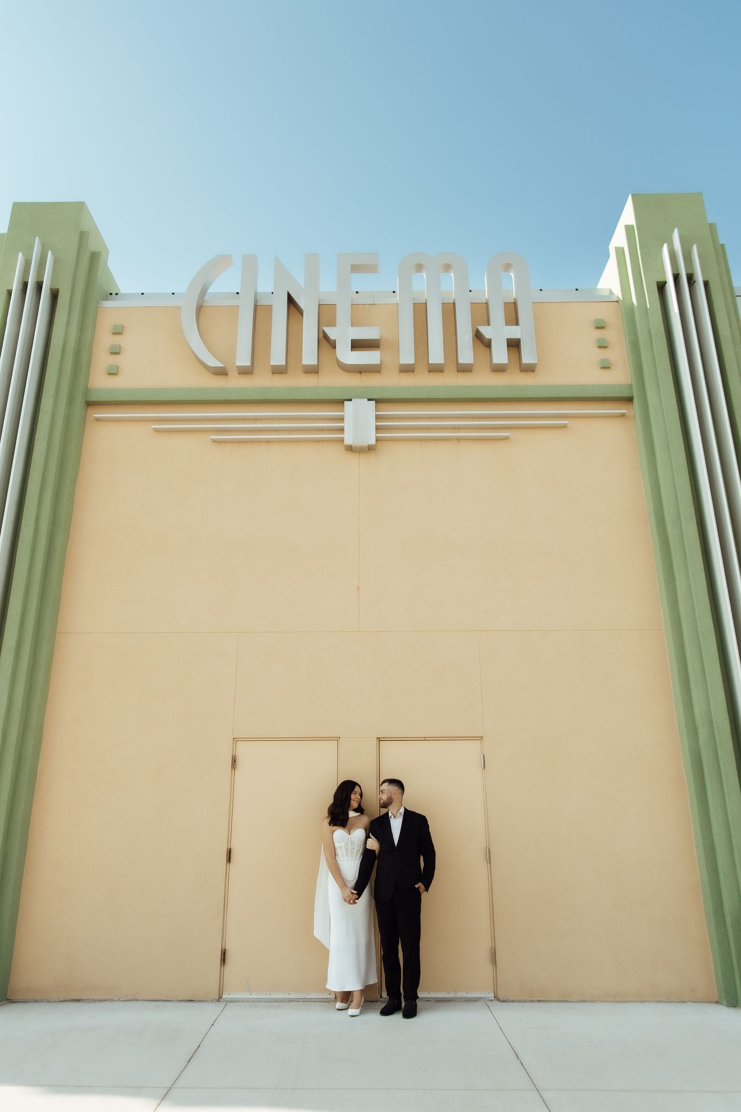 Couple in formal attire standing before an Art Deco cinema facade under a clear blue sky.
