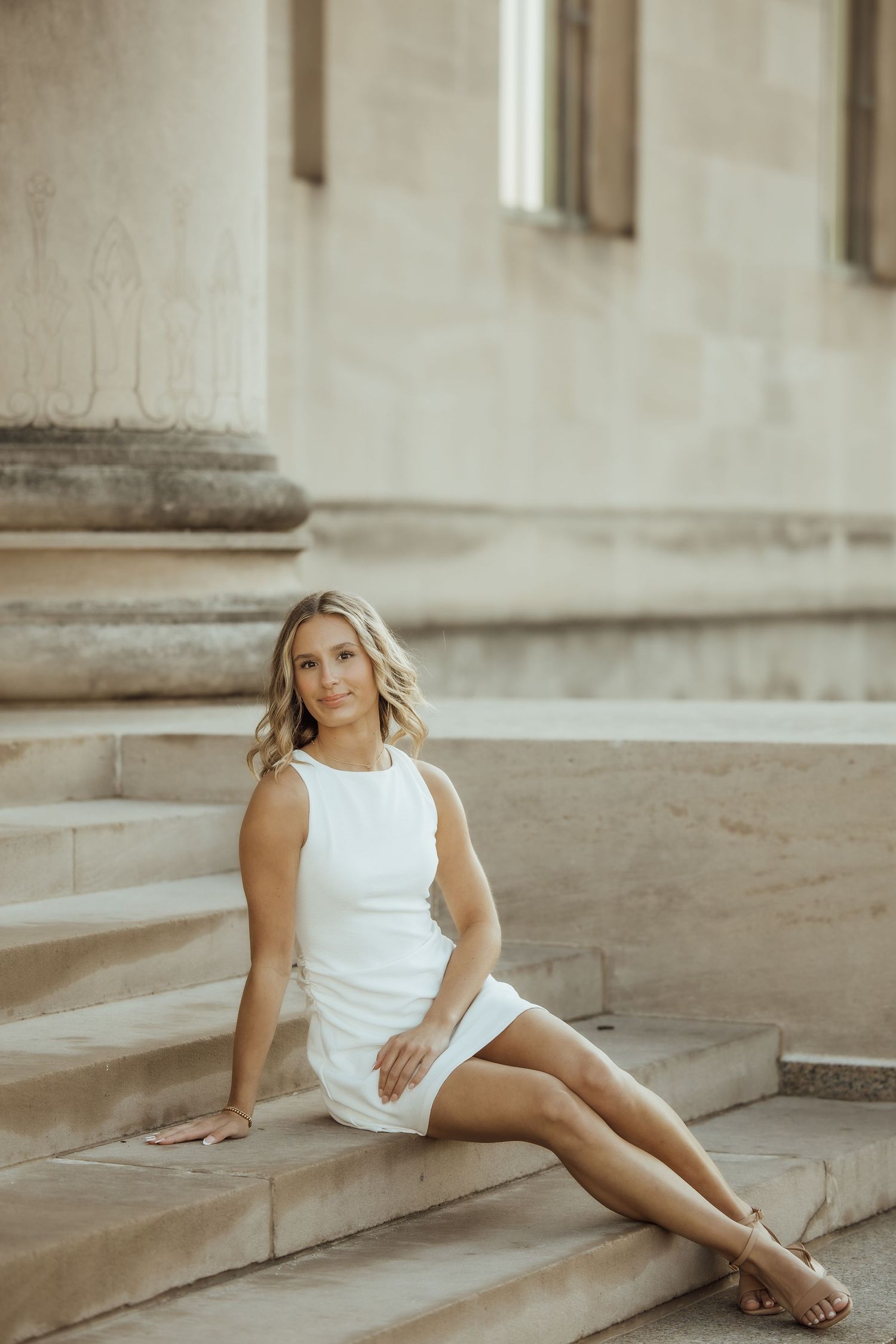 Blonde woman in white sleeveless dress sits gracefully on stone steps near columns.