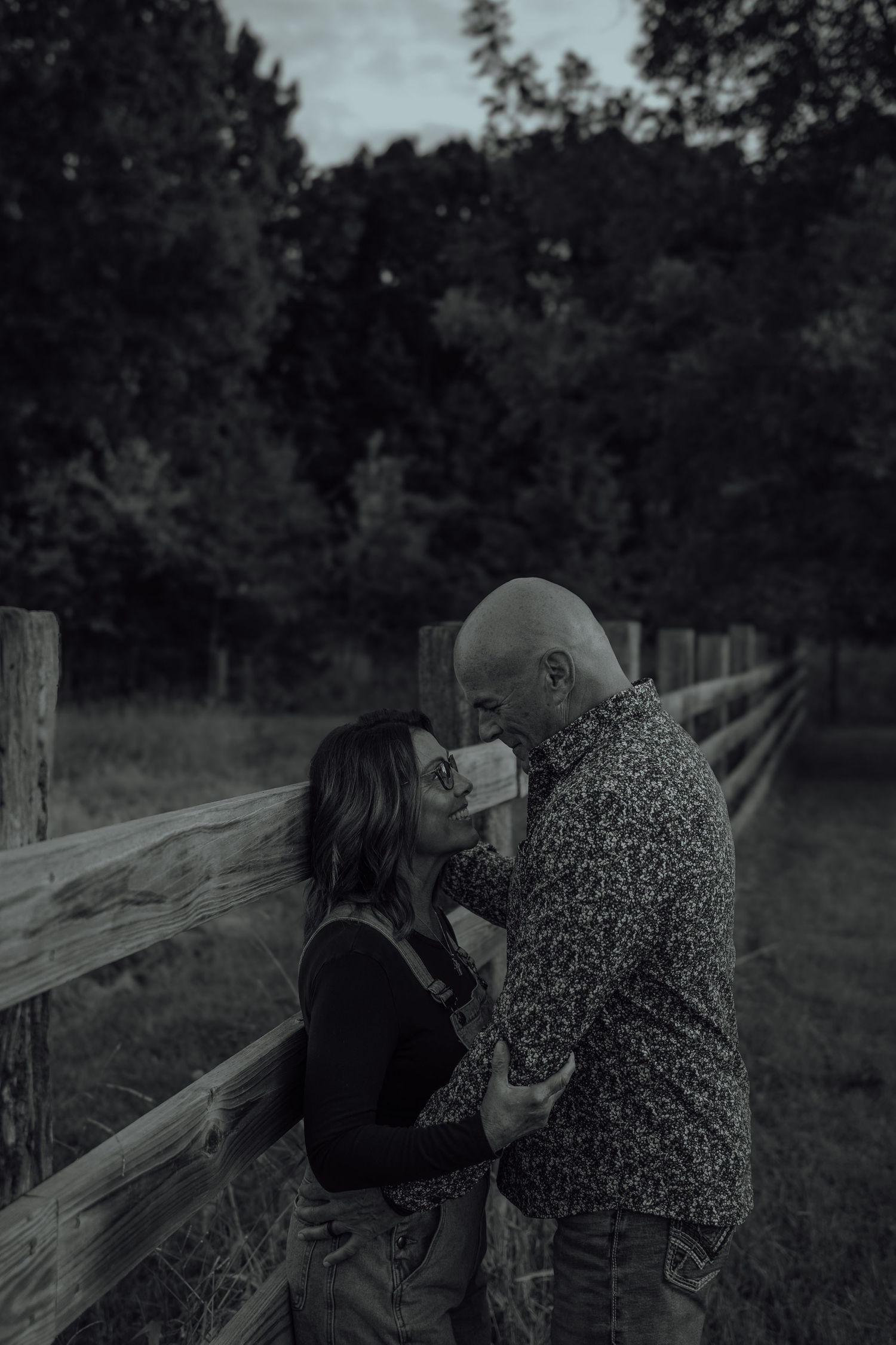 Black and white romantic couple almost kissing by wooden fence at dusk in wooded countryside setting.
