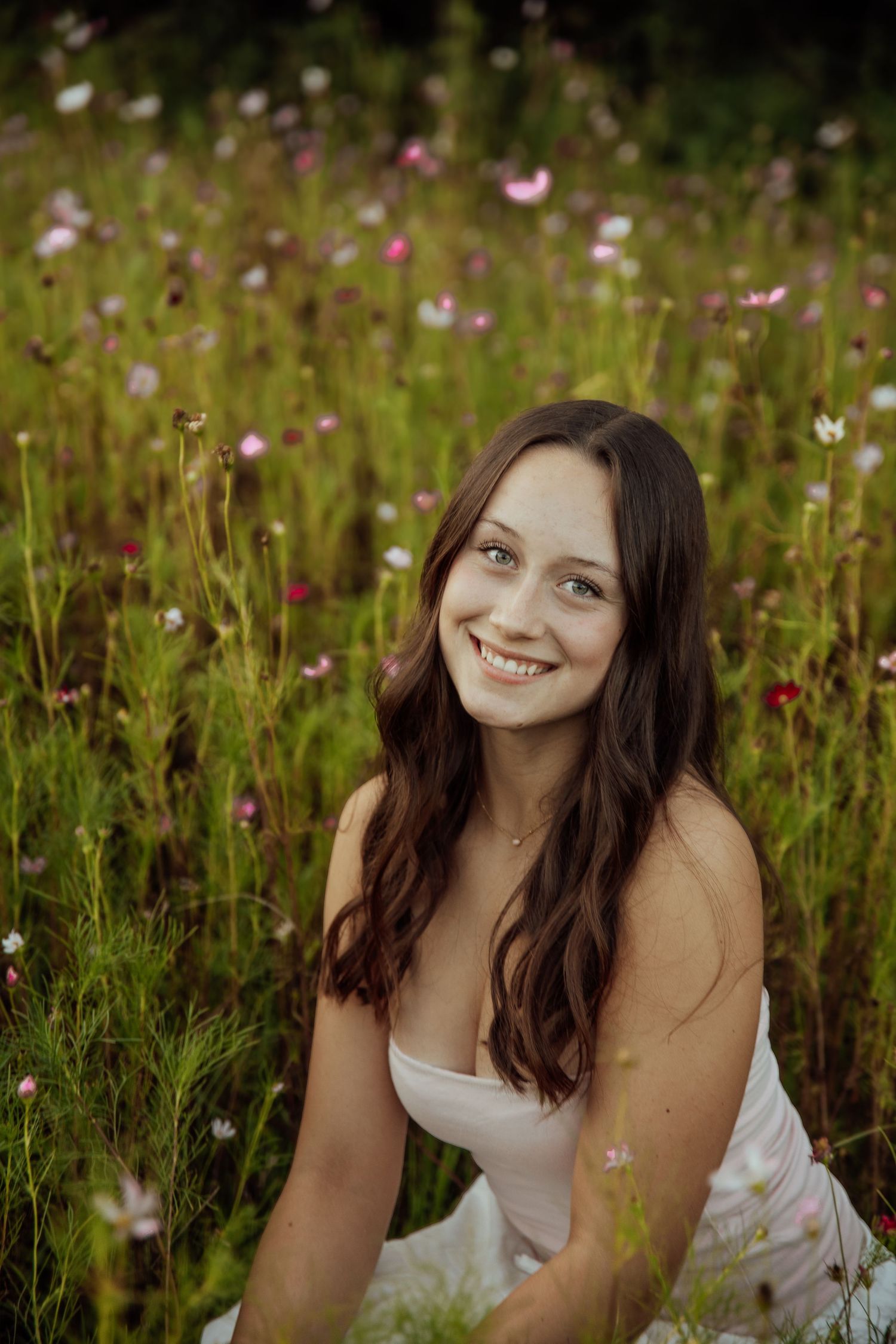 Smiling brunette woman in white dress sitting in colorful wildflower meadow with pink blooms surrounding her.