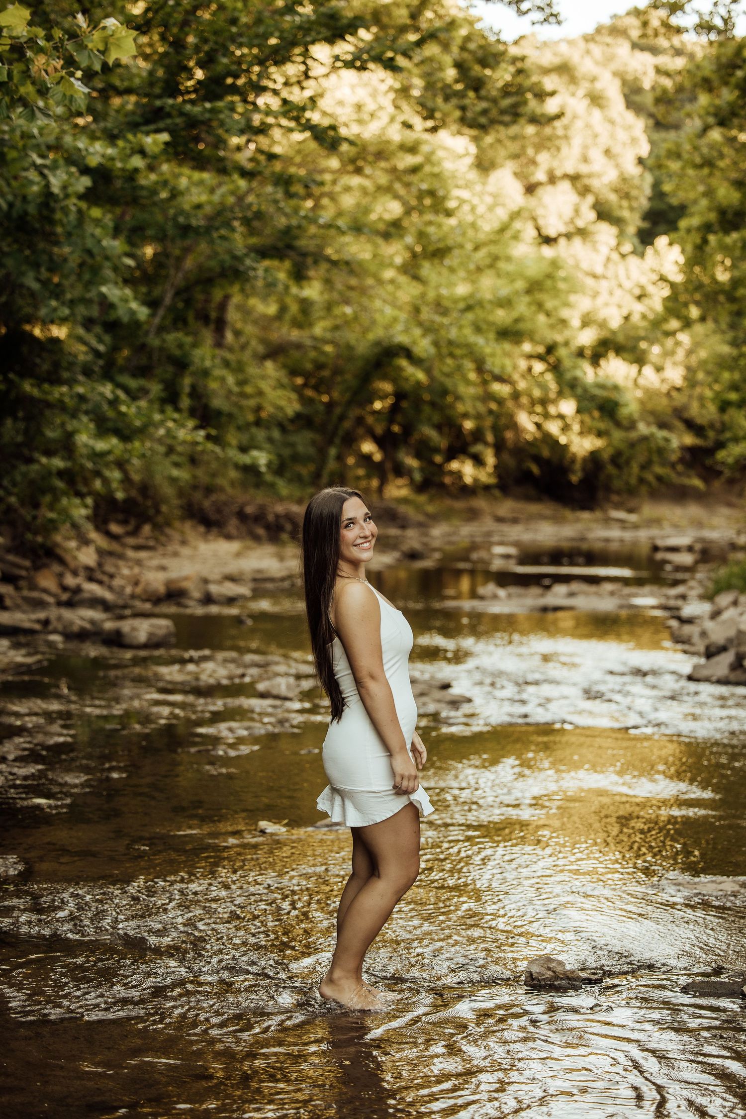 Young woman in white dress standing barefoot in a shallow forest creek, smiling over her shoulder.
