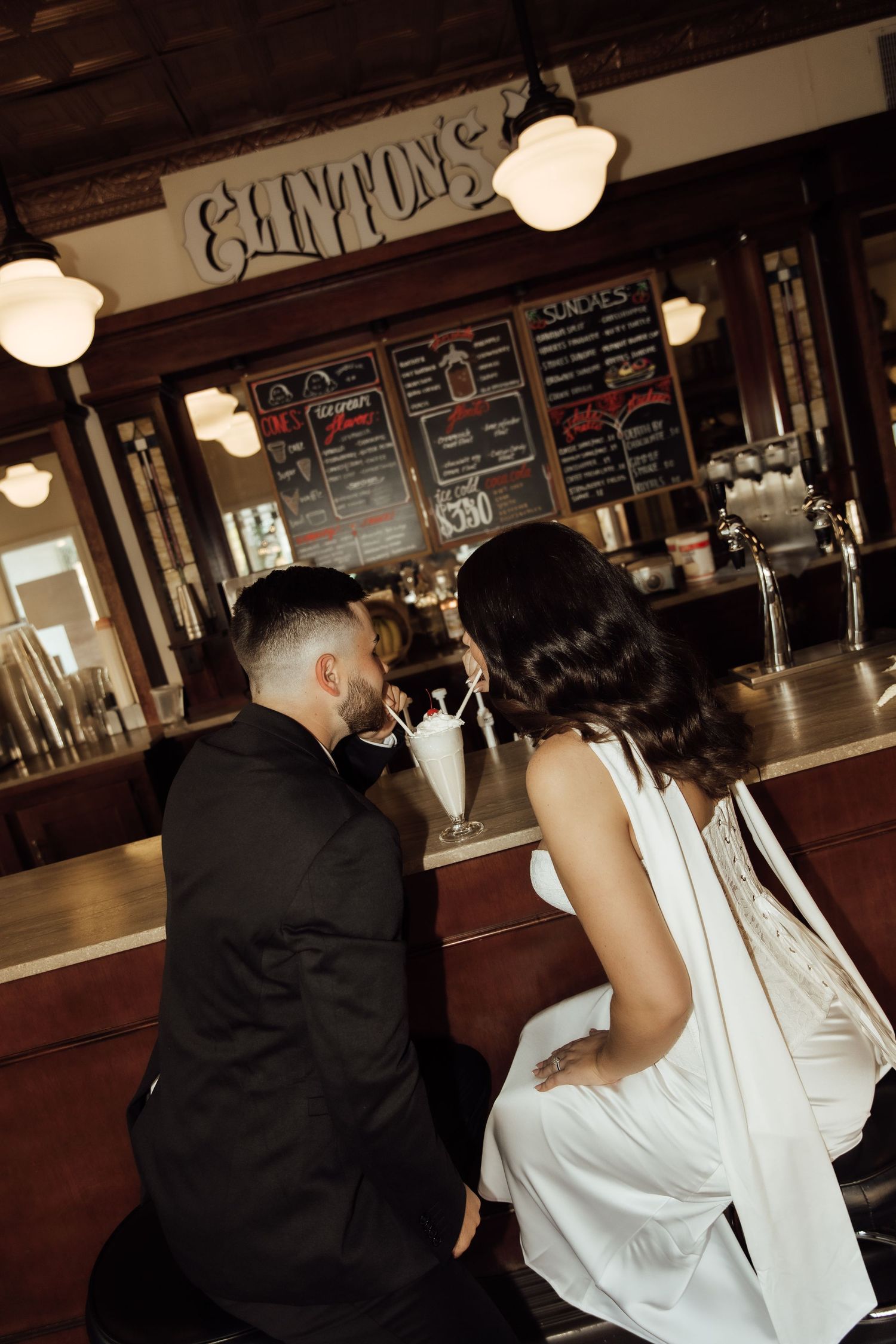 Couple in wedding attire sharing a milkshake at a vintage diner counter, romantic date scene.