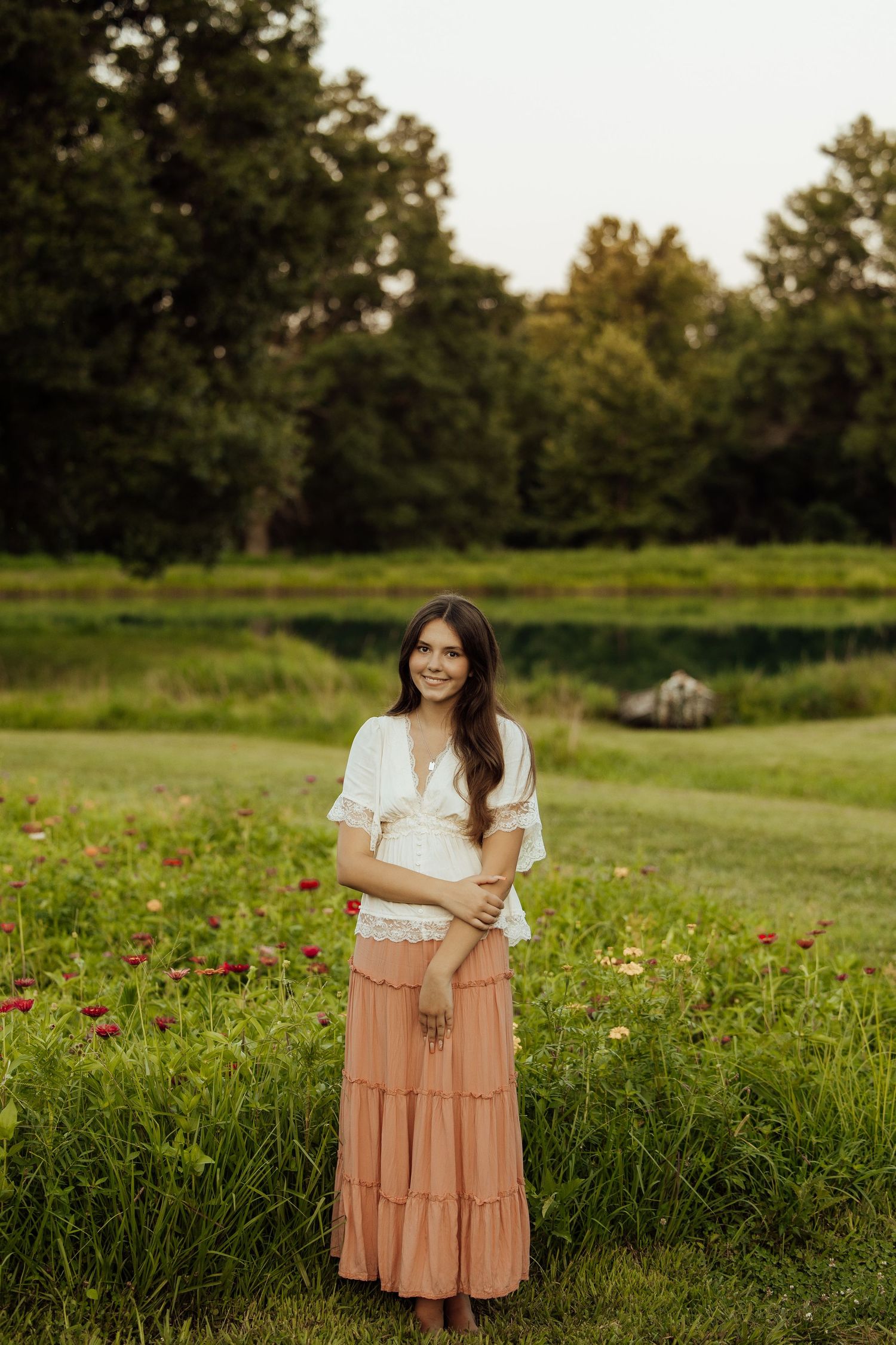 Young woman in white lace top and pink maxi skirt smiling in a lush green meadow with wildflowers.