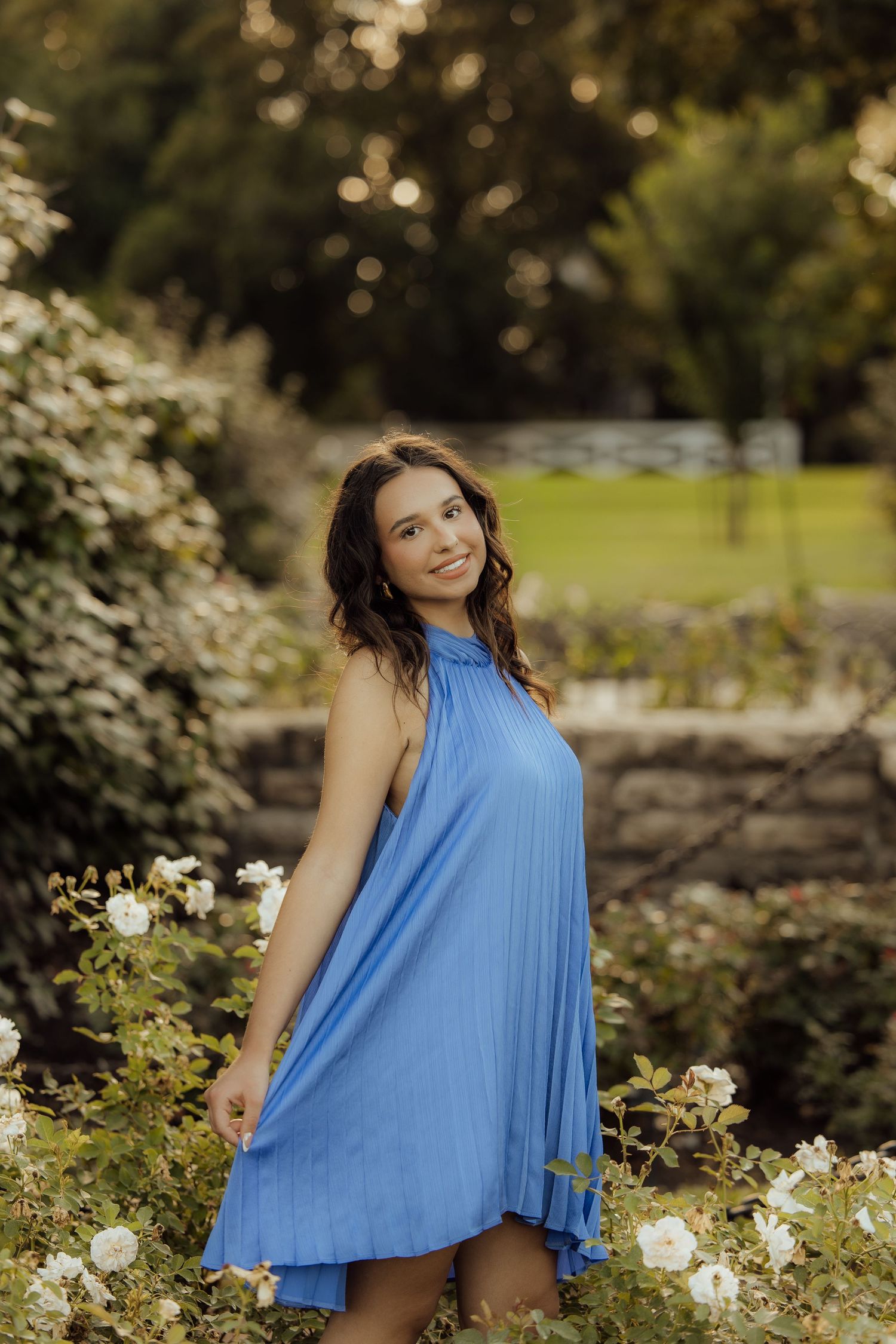 Young woman in blue pleated halter dress smiling among white roses in a lush garden setting.