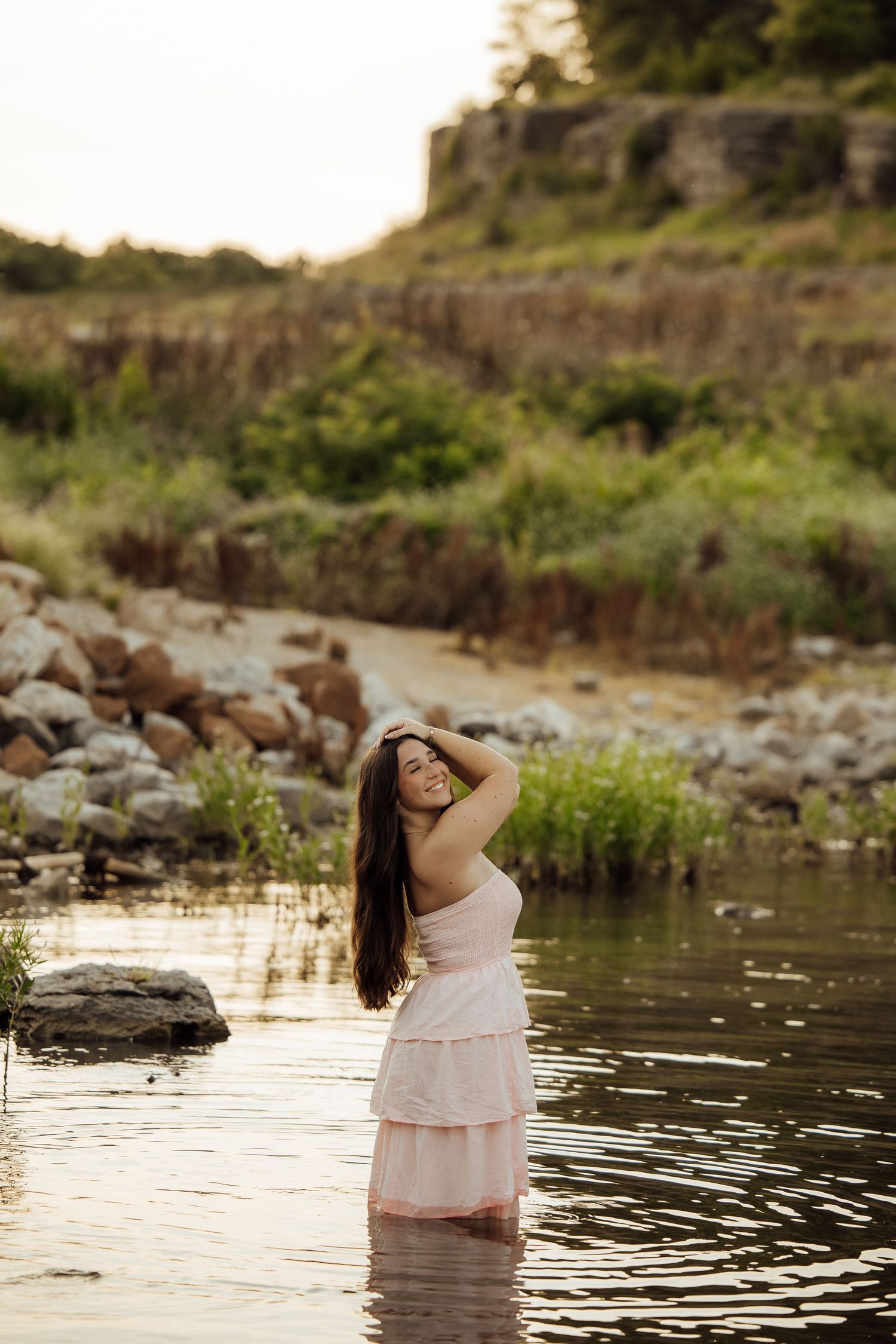 Young woman in pink dress standing in shallow river water at golden hour, surrounded by rocks and greenery.