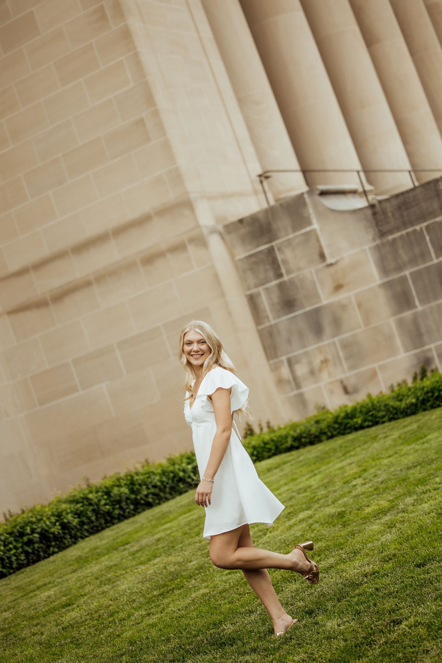 Elegant blonde woman in white flutter-sleeve dress walking on green lawn beside stone building.