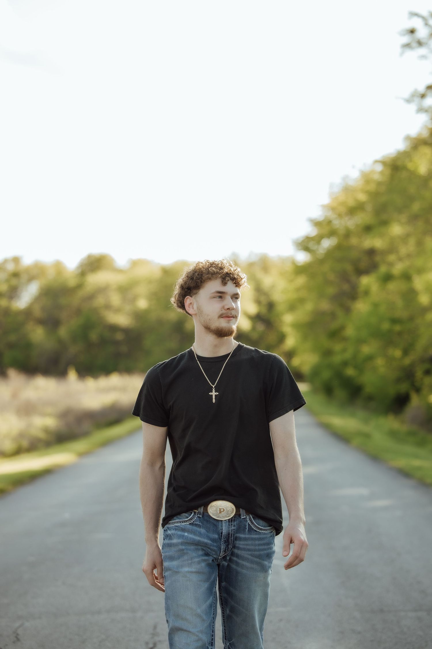 Young man in black t-shirt and jeans walks down a tree-lined road in natural light.