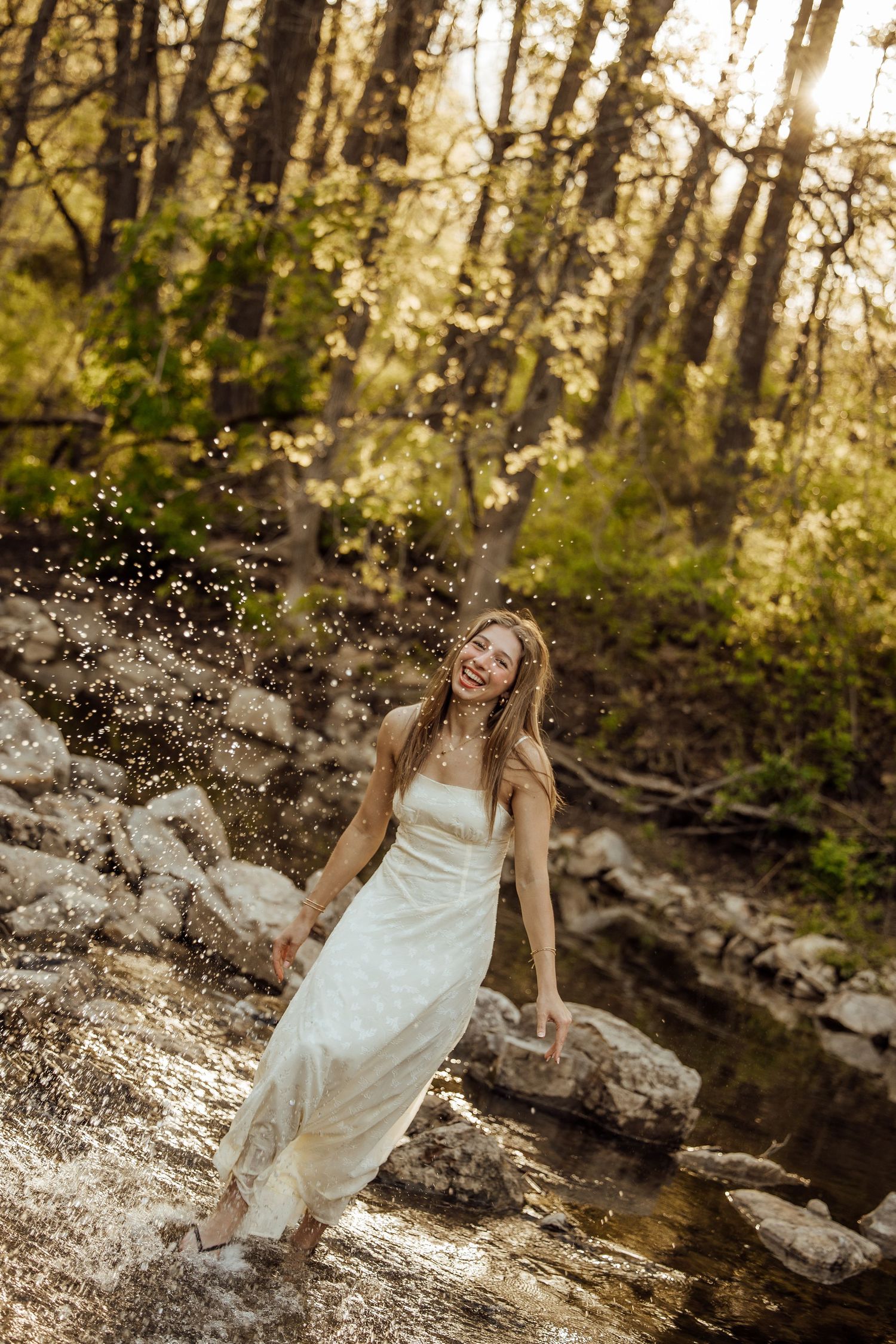 Joyful woman in white dress splashing water in a sunlit rocky creek in the woods.