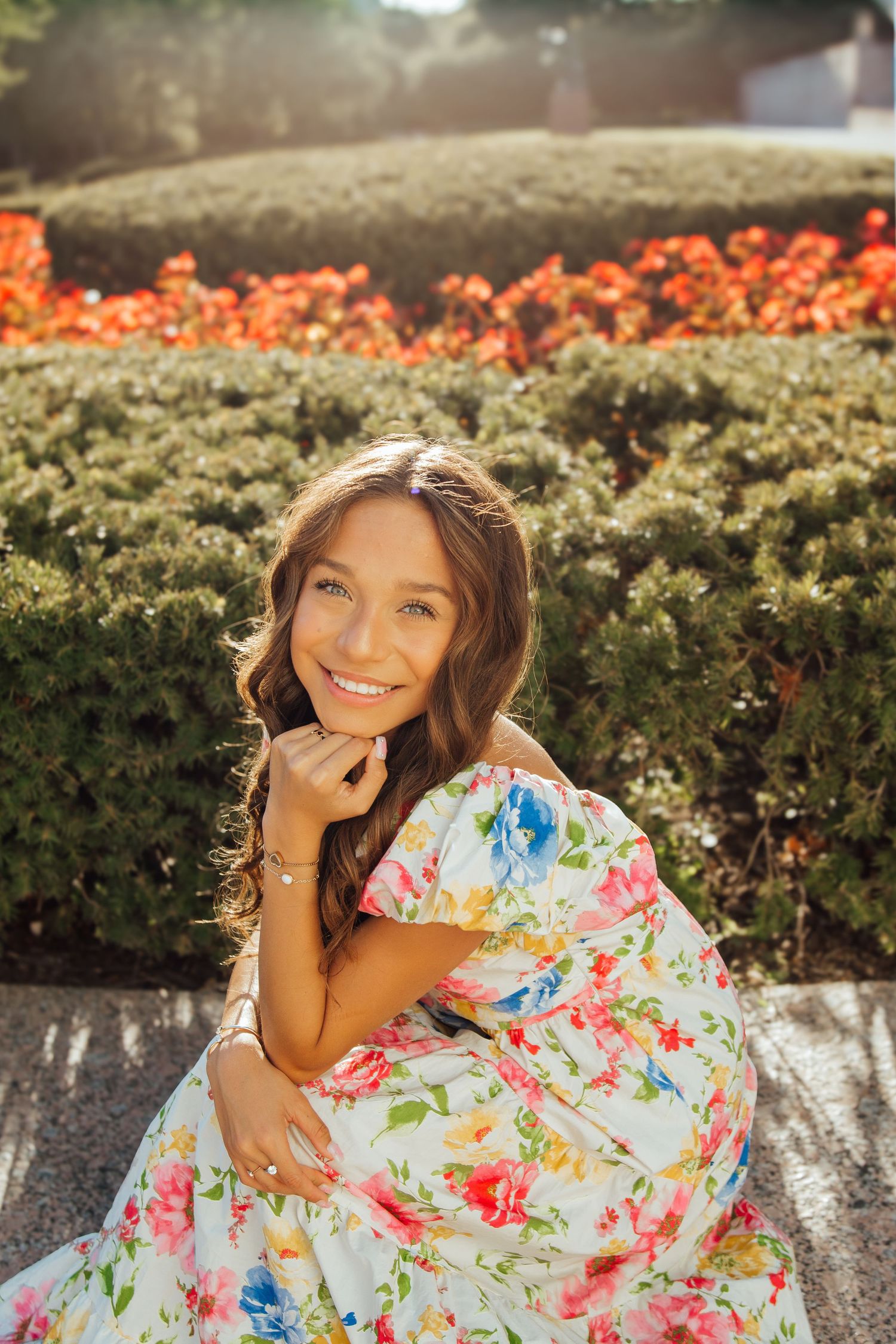 Smiling young woman in floral dress sitting outdoors near red flowers and green hedges in sunlight.
