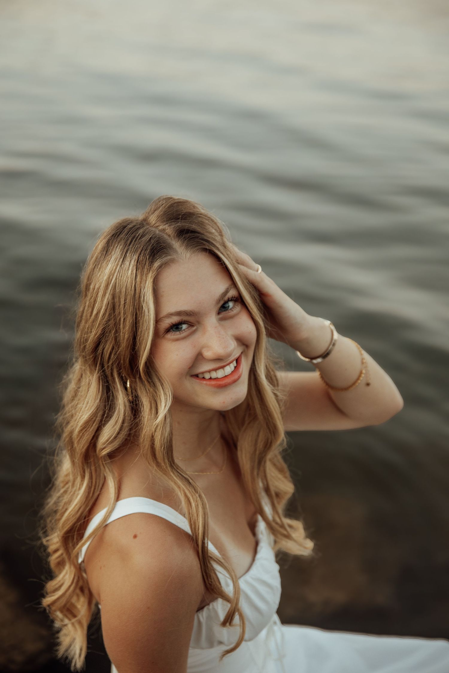 Smiling blonde woman in white dress with hand in hair posing near shimmering water at dusk.