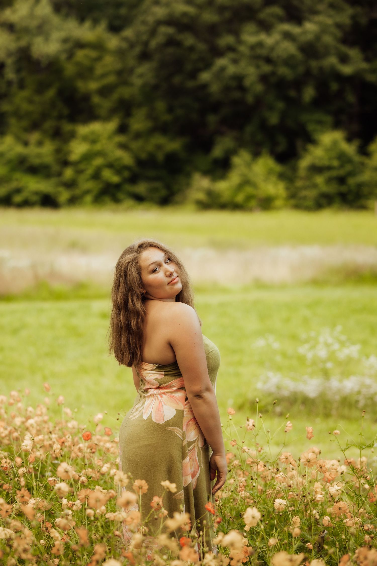 Young woman in floral dress standing in a wildflower meadow with lush green trees in the background.