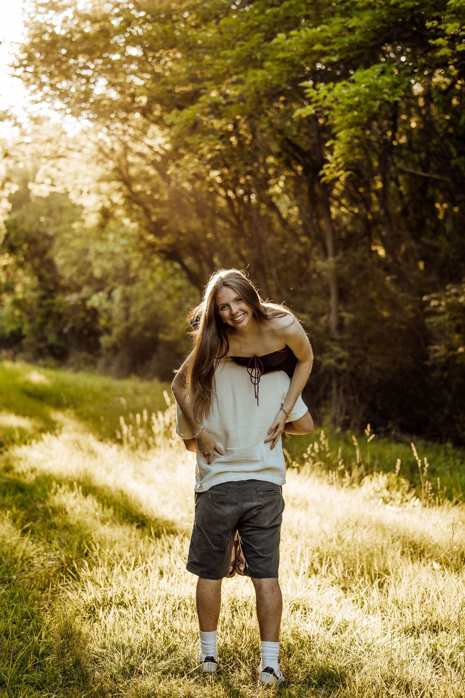 Couple laughing outdoors, woman piggyback riding man in sunny field with green trees in background.