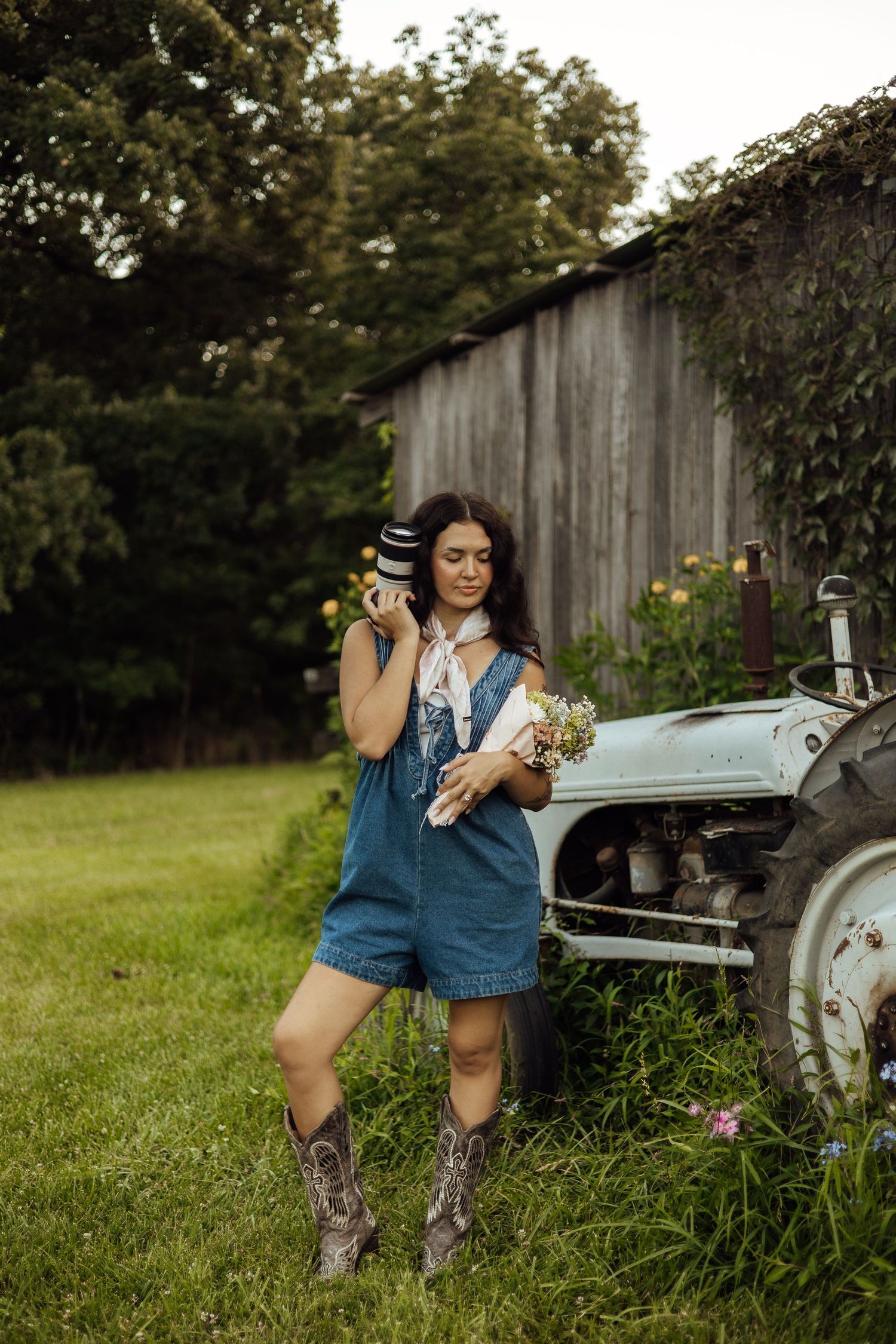 Woman in denim overalls and cowboy boots holds coffee cup near vintage tractor on a rustic farm.