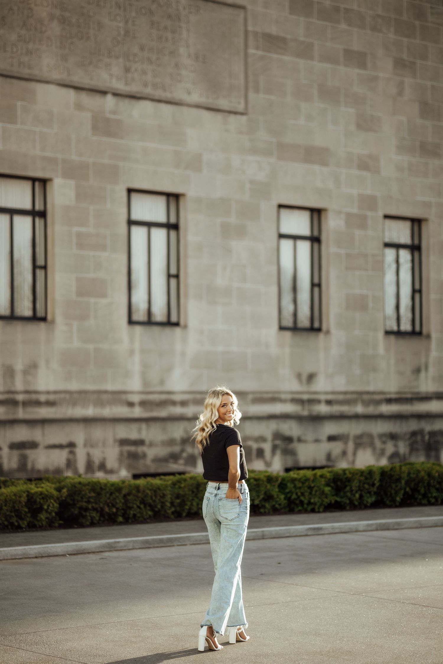 Blonde woman in black top and light blue jeans posing outdoors against a stone building wall.