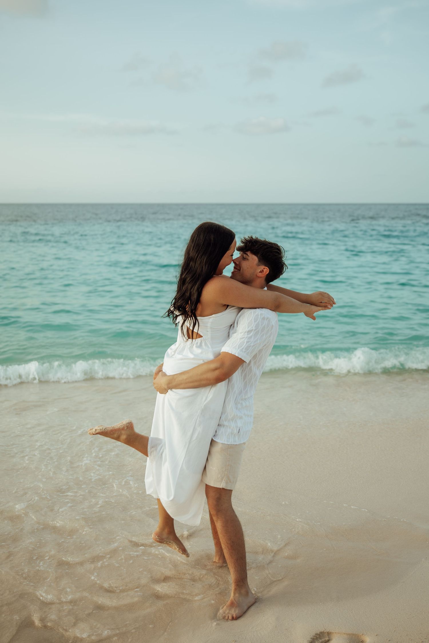 Happy couple embracing on a tropical beach, man lifting woman in white dress near turquoise ocean waves.