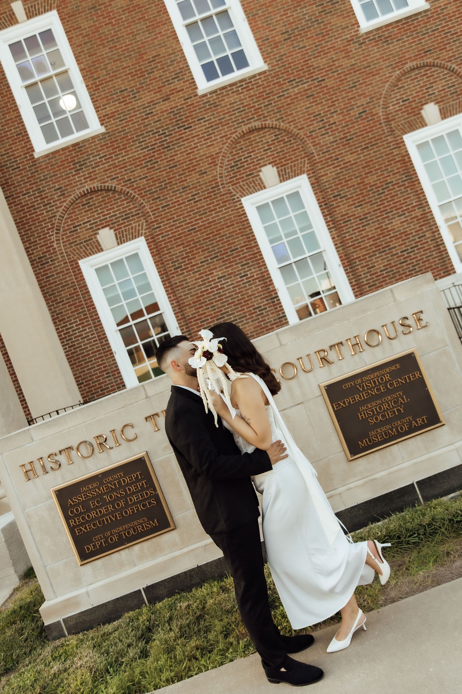 Newlywed couple sharing a romantic kiss in front of a Historic Courthouse, bride in white dress holding bouquet.