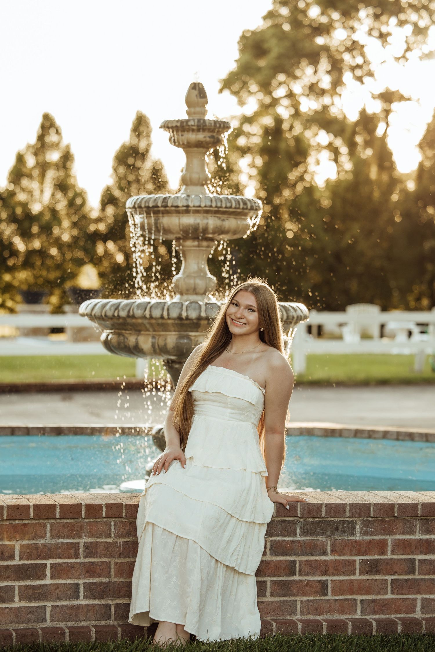 Young woman in white strapless dress posing by ornate stone fountain at golden hour in lush garden.
