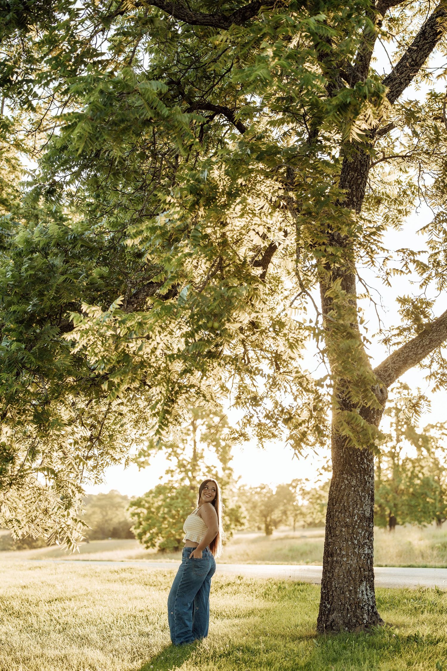 Young woman in jeans and crop top standing under a large green tree in a sunlit park during golden hour.