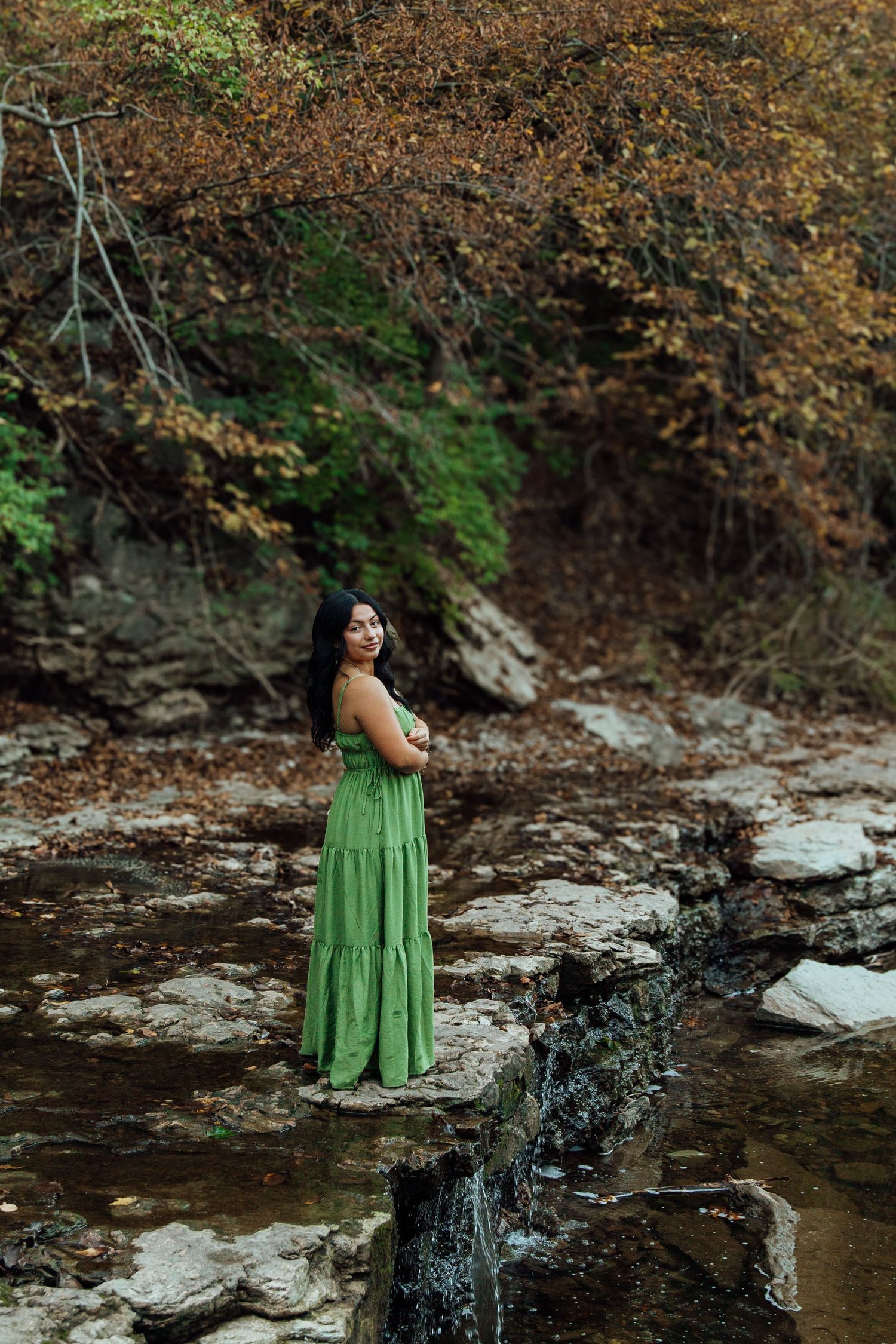 Woman in green maxi dress standing on rocks by a small waterfall in an autumn forest.