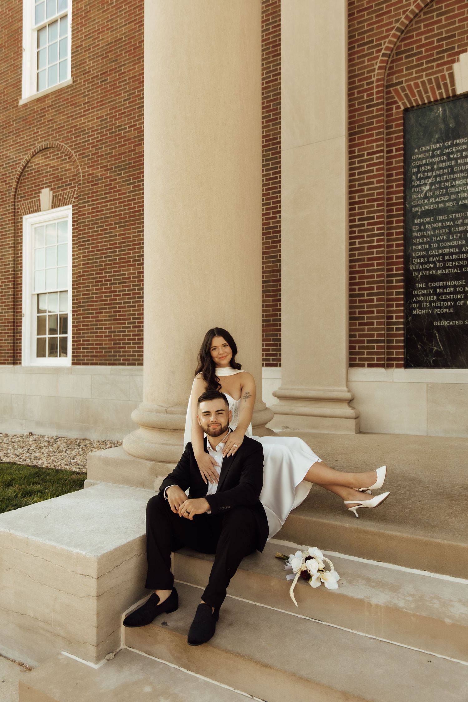 Bride and groom posing on brick building steps, she in white dress, he in black suit, with bridal bouquet nearby.