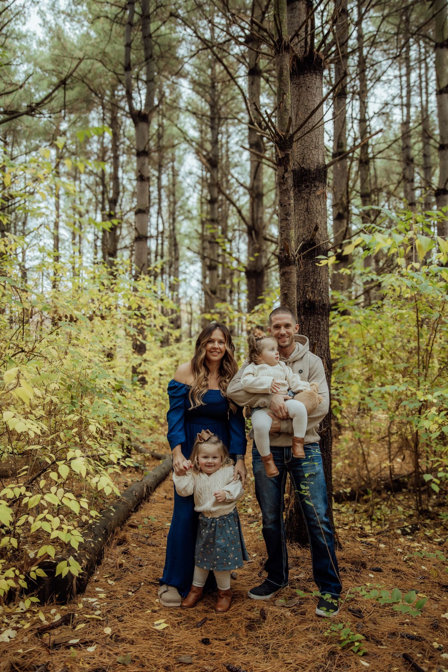 Young family of four posing among tall pine trees during an autumn outdoor family photography session.