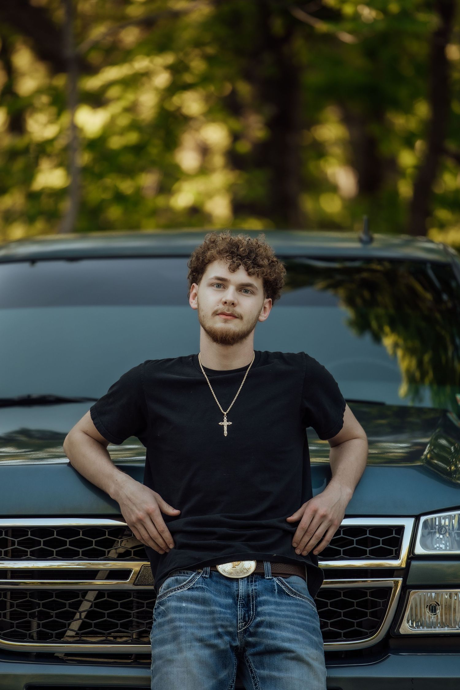 Young man with curly hair in black t-shirt leans against dark truck in wooded setting.