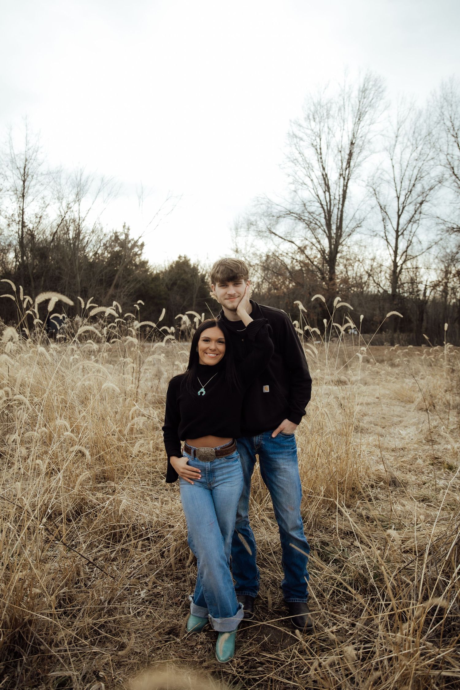 Young couple embracing in a golden dried grass field with bare winter trees in the background.