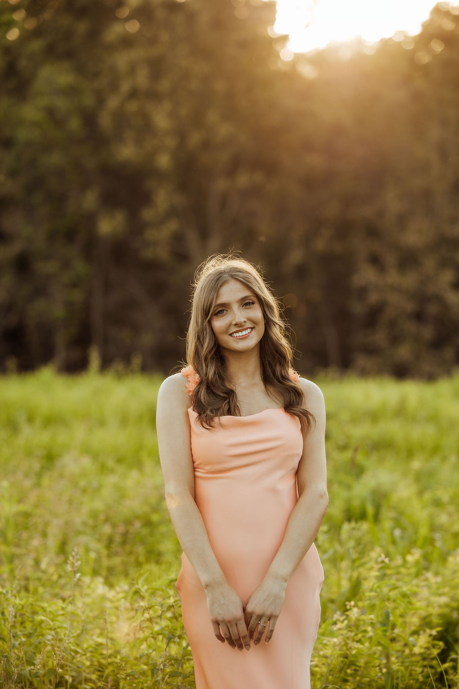 Smiling young woman in peach slip dress standing in lush green field at sunset.