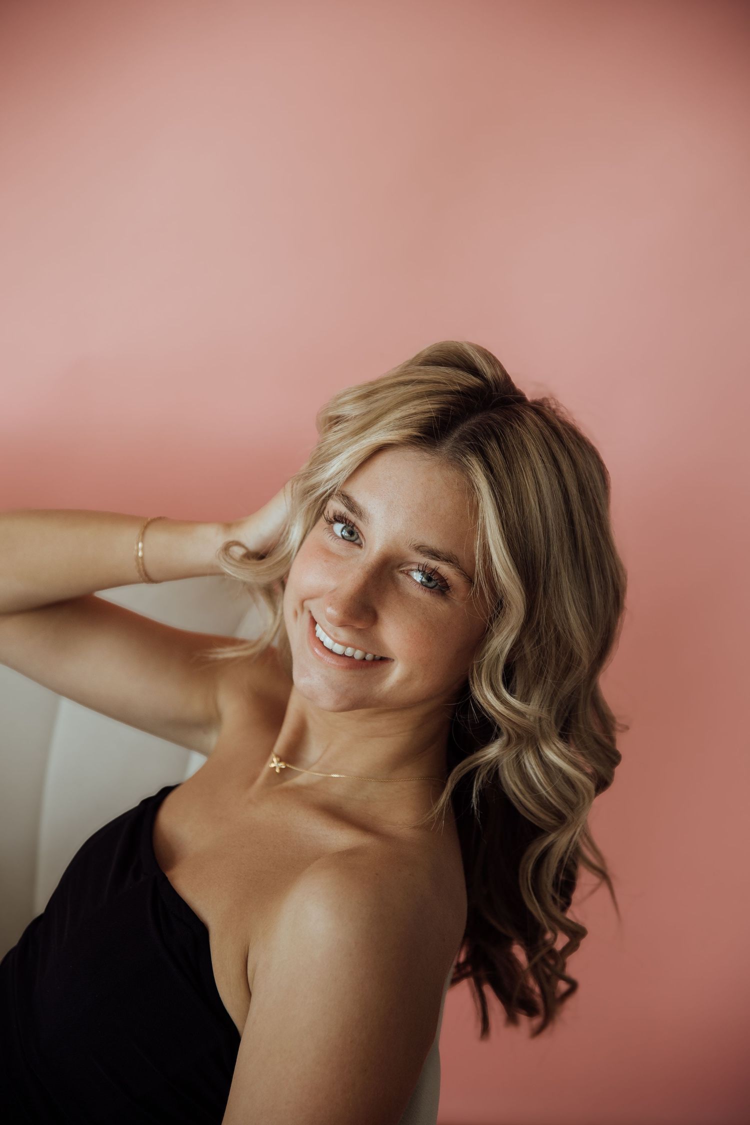 Smiling blonde woman in black top with gold necklace posing against a soft pink background.