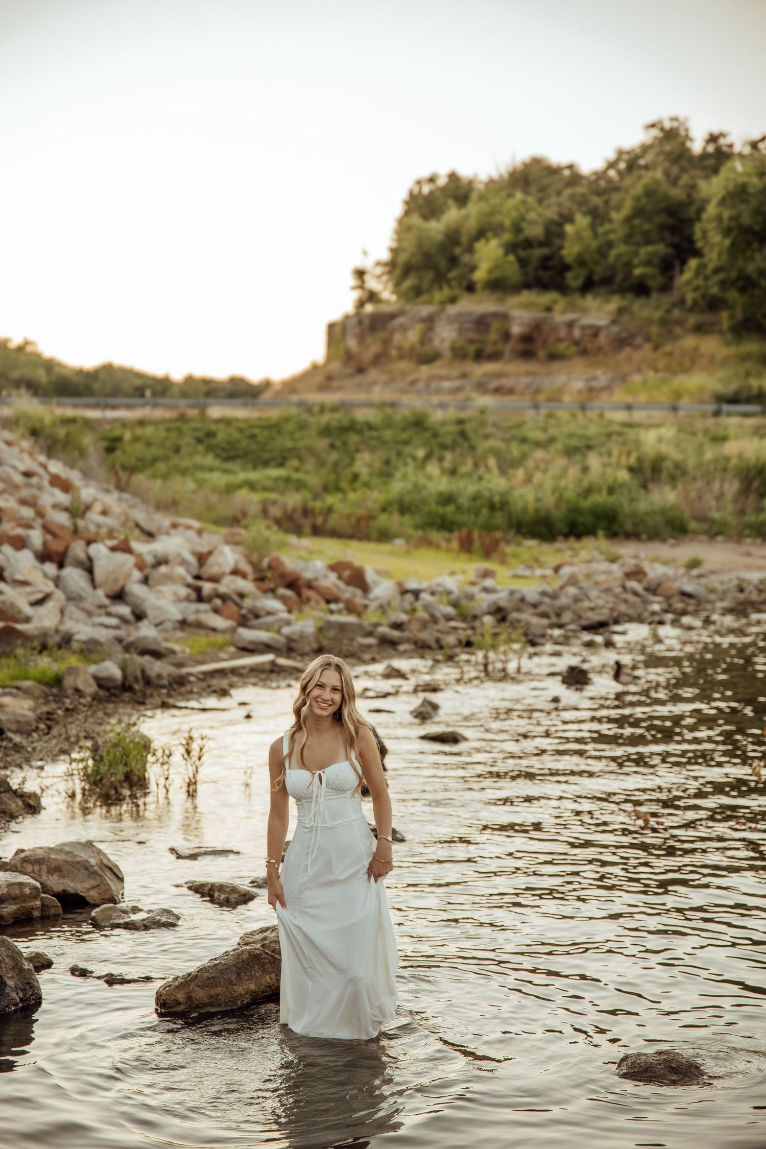 Blonde woman in white dress standing in shallow rocky river with scenic landscape at golden hour.