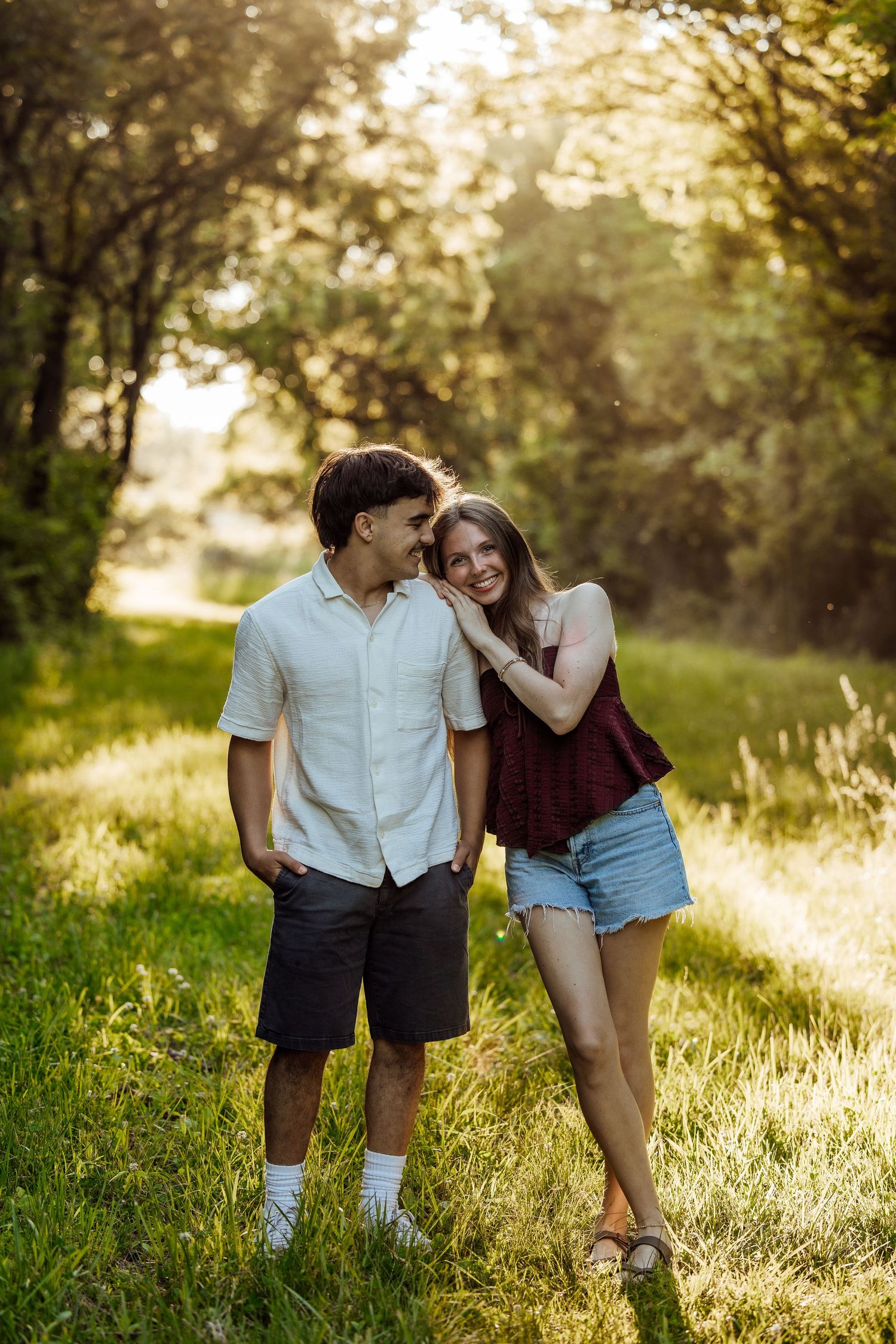 A young couple smiles and embraces on a sunlit grassy path surrounded by lush green trees.