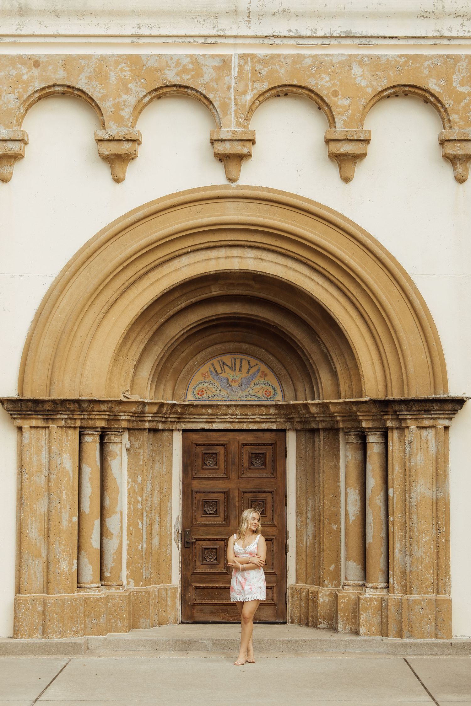 Young blonde woman in floral dress standing before ornate arched church entrance with decorative stonework.
