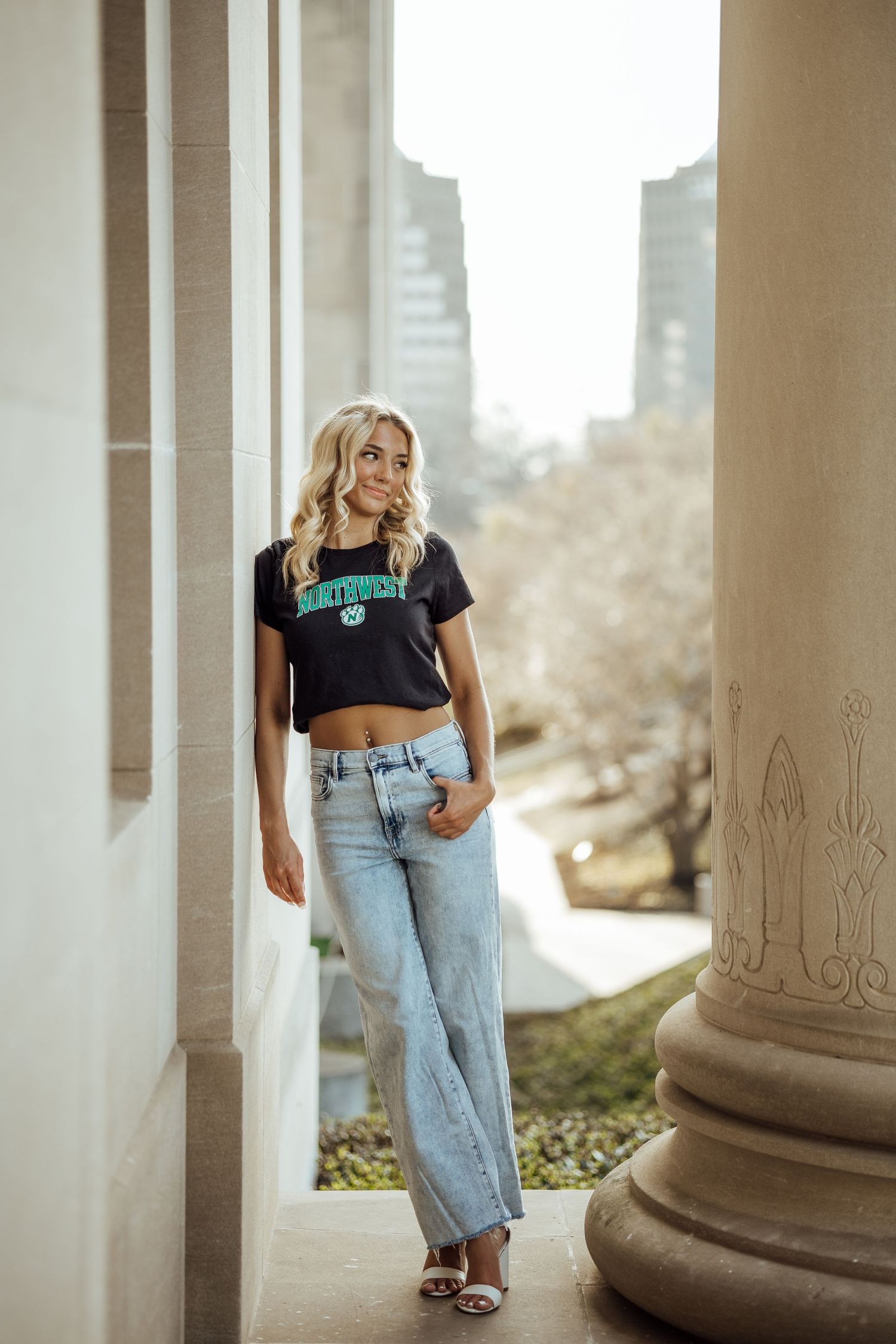 Blonde woman in black North Dakota crop top and jeans poses between stone columns outdoors.