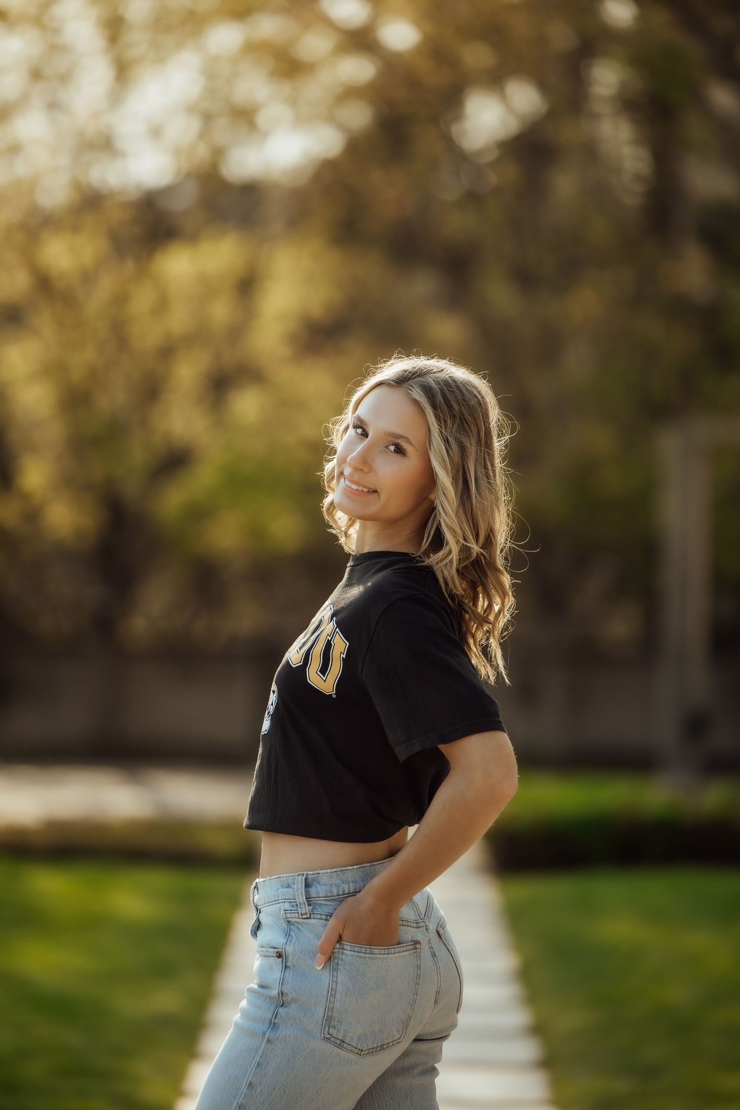 Young blonde woman in black crop top and jeans poses on sunlit outdoor pathway.