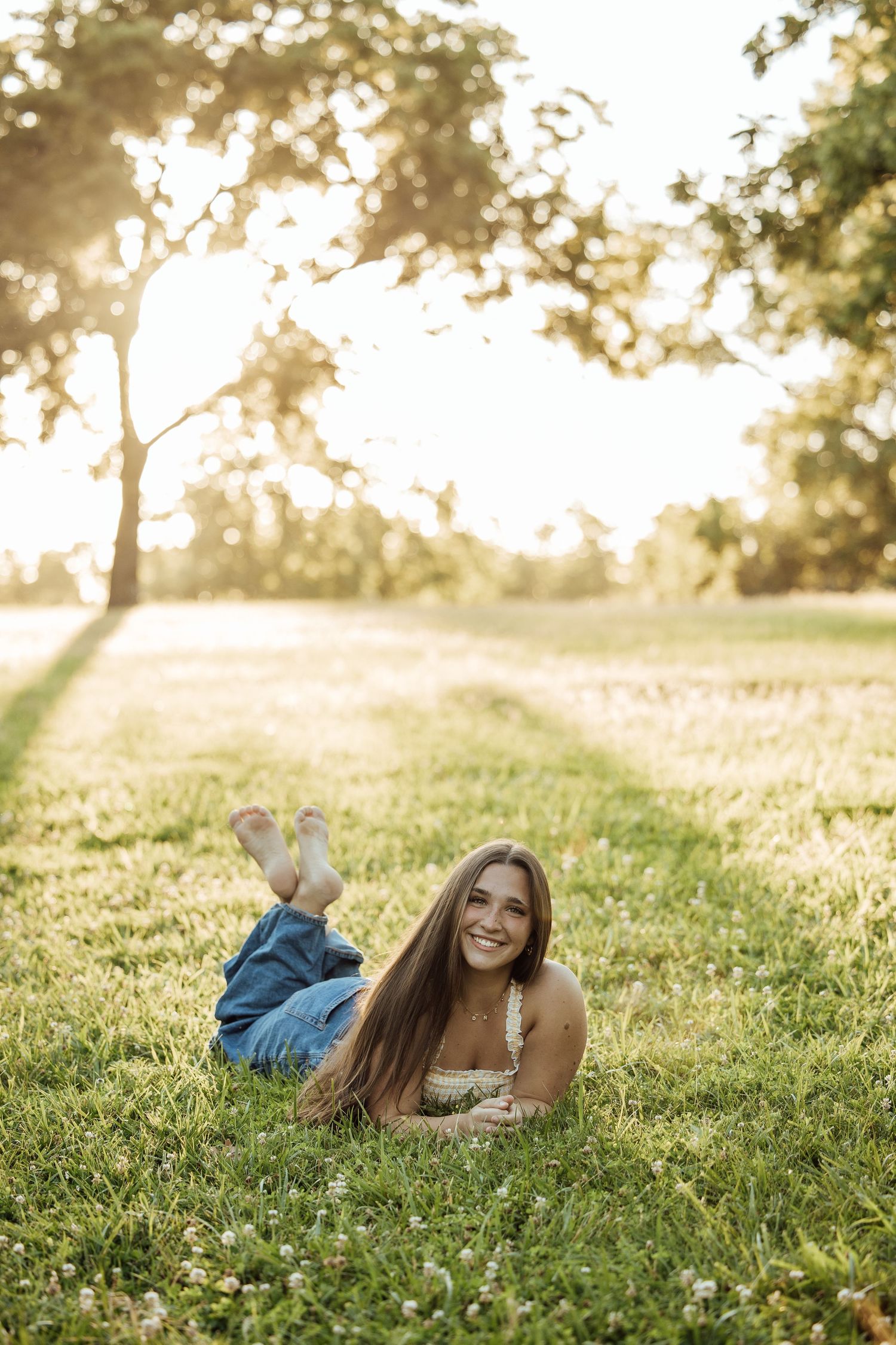 Young woman lying on green grass in a sunny park, smiling, wearing jeans and a floral top at golden hour.