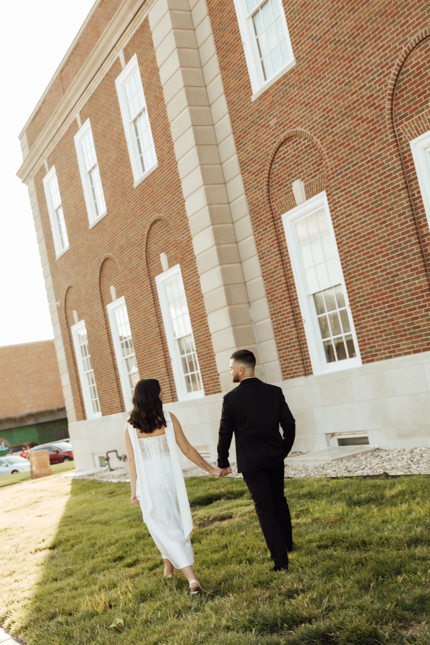 A couple in wedding attire holds hands while walking on grass beside a red brick building.