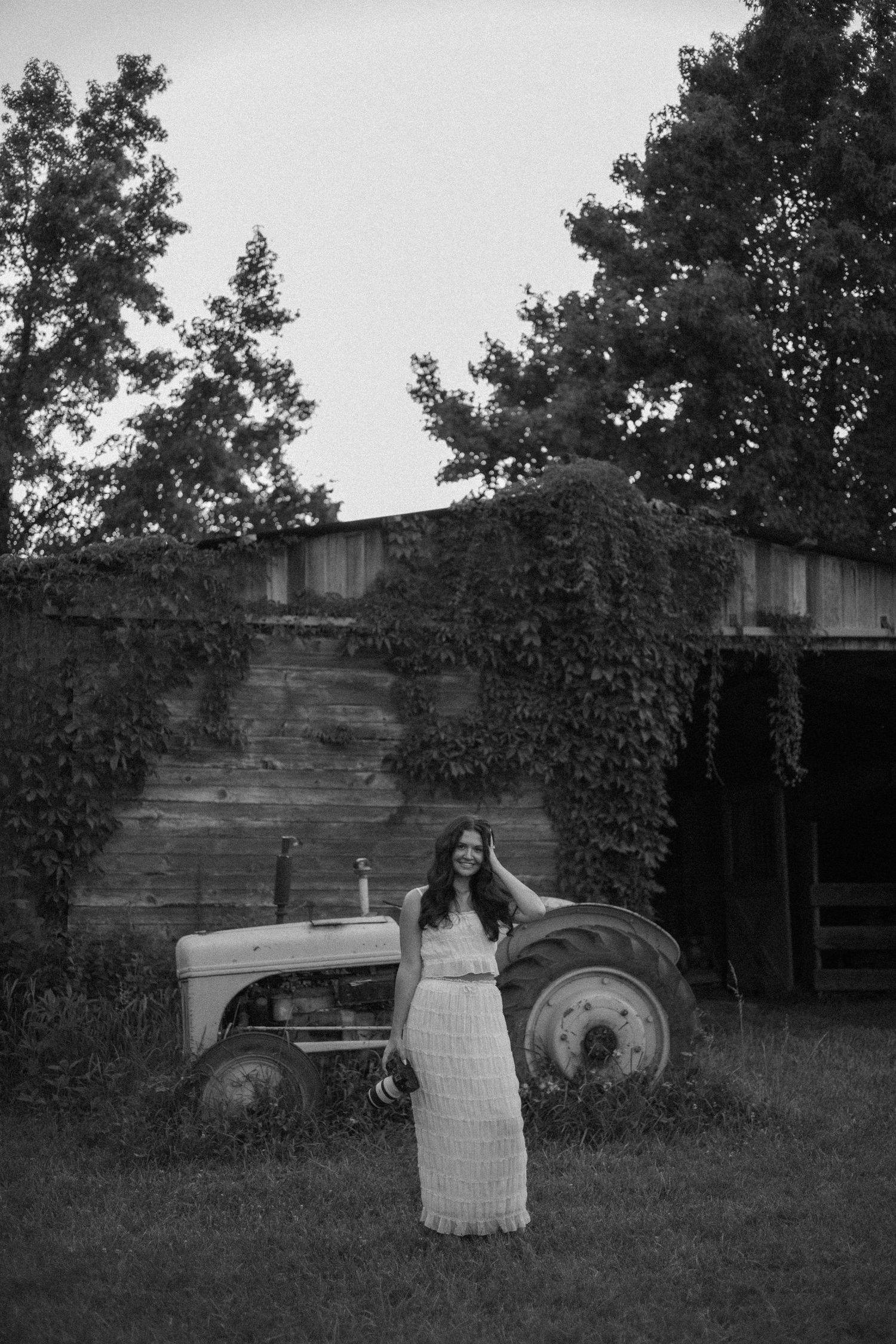Black and white photo of a young woman in a lace dress posing beside a vintage tractor near an ivy-covered barn.