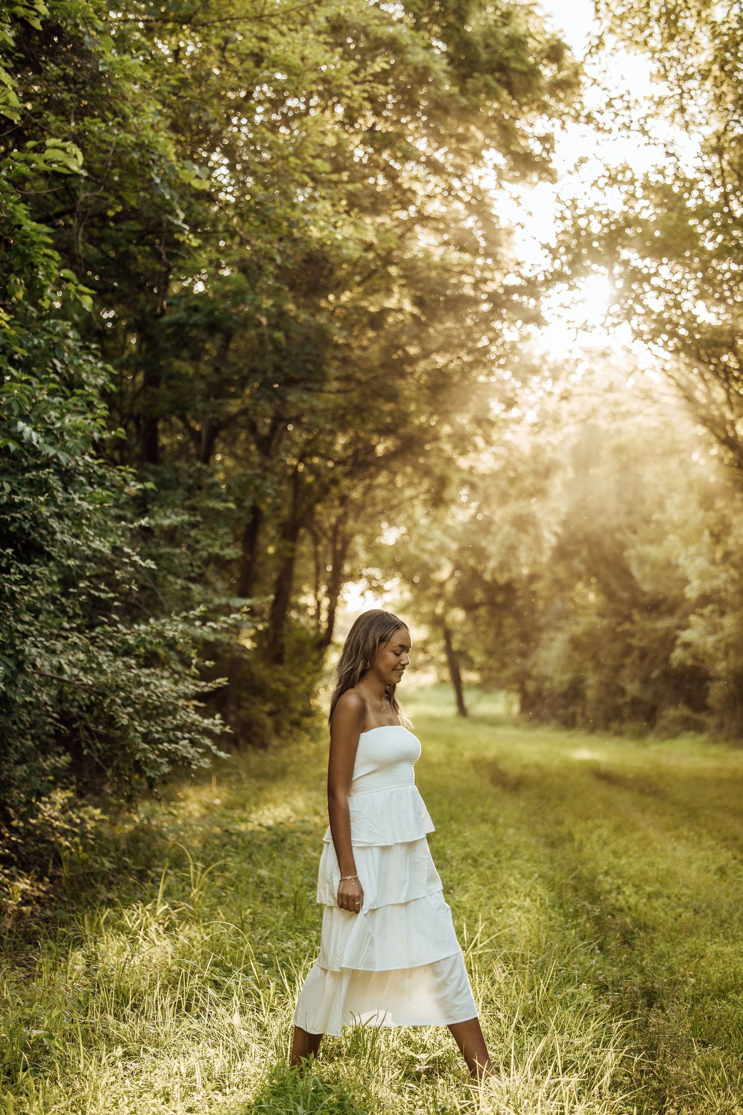 Young woman in white tiered strapless dress standing on a sunlit forest path surrounded by lush green trees.