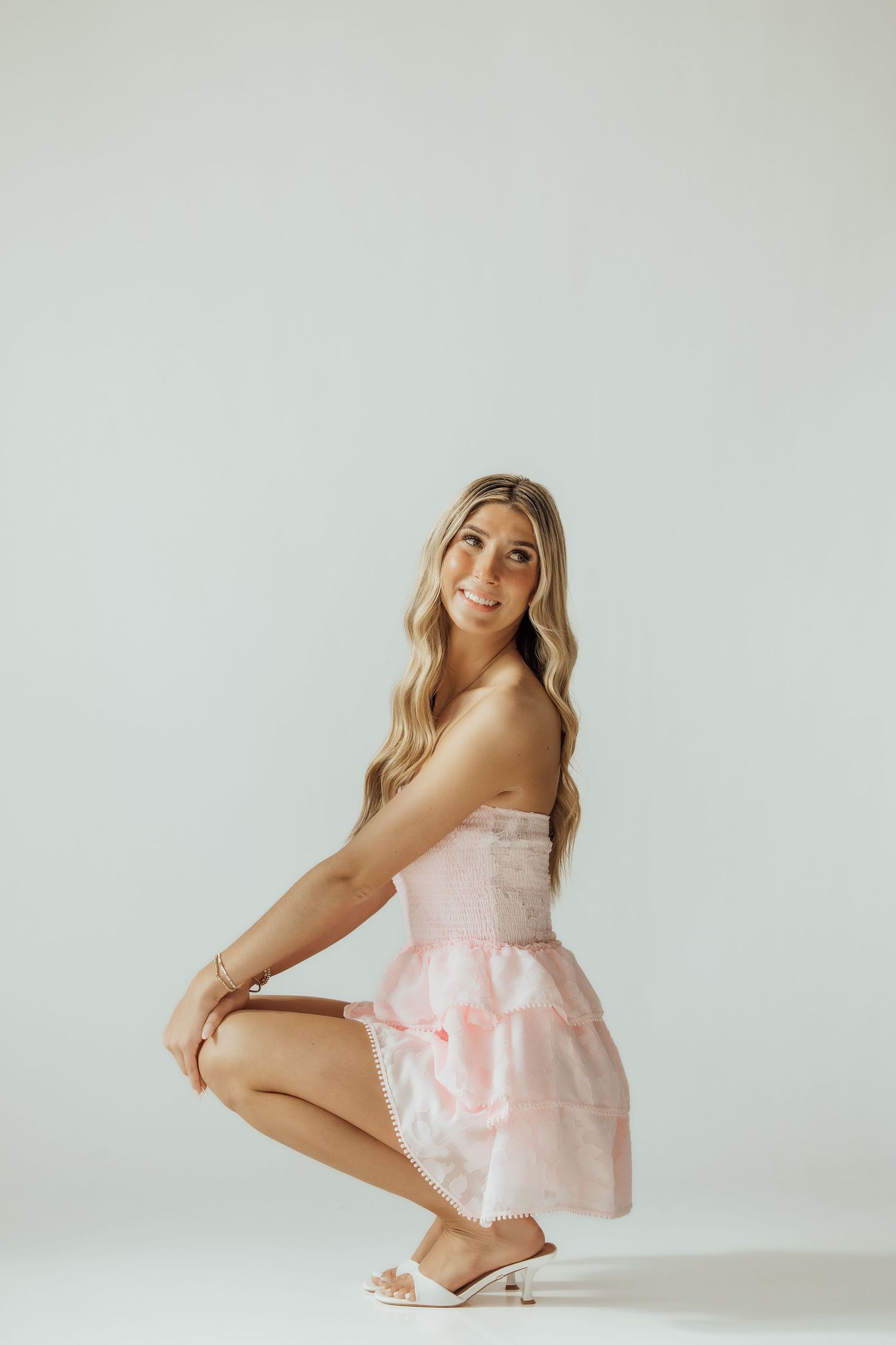 Young blonde woman in pink strapless mini dress crouching and smiling against white studio background.