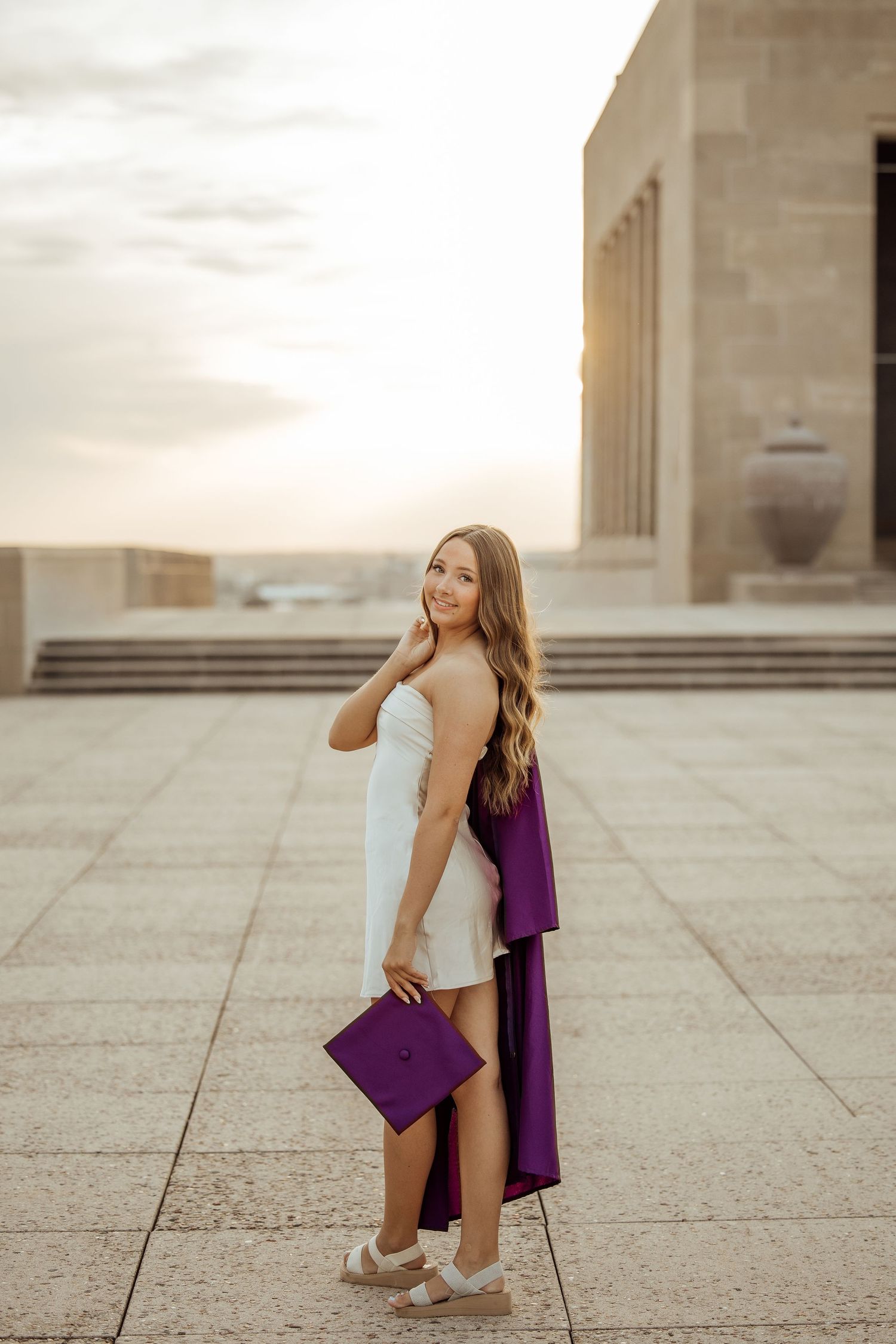Beautiful graduate in white dress holding purple cap and gown at sunset on university plaza.