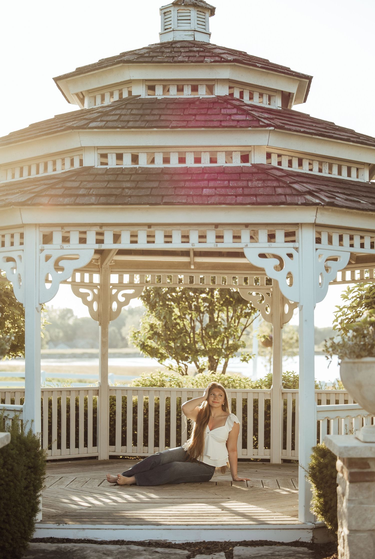 Woman sitting gracefully inside a white Victorian gazebo with ornate details, bathed in warm golden sunlight near a serene waterfront.