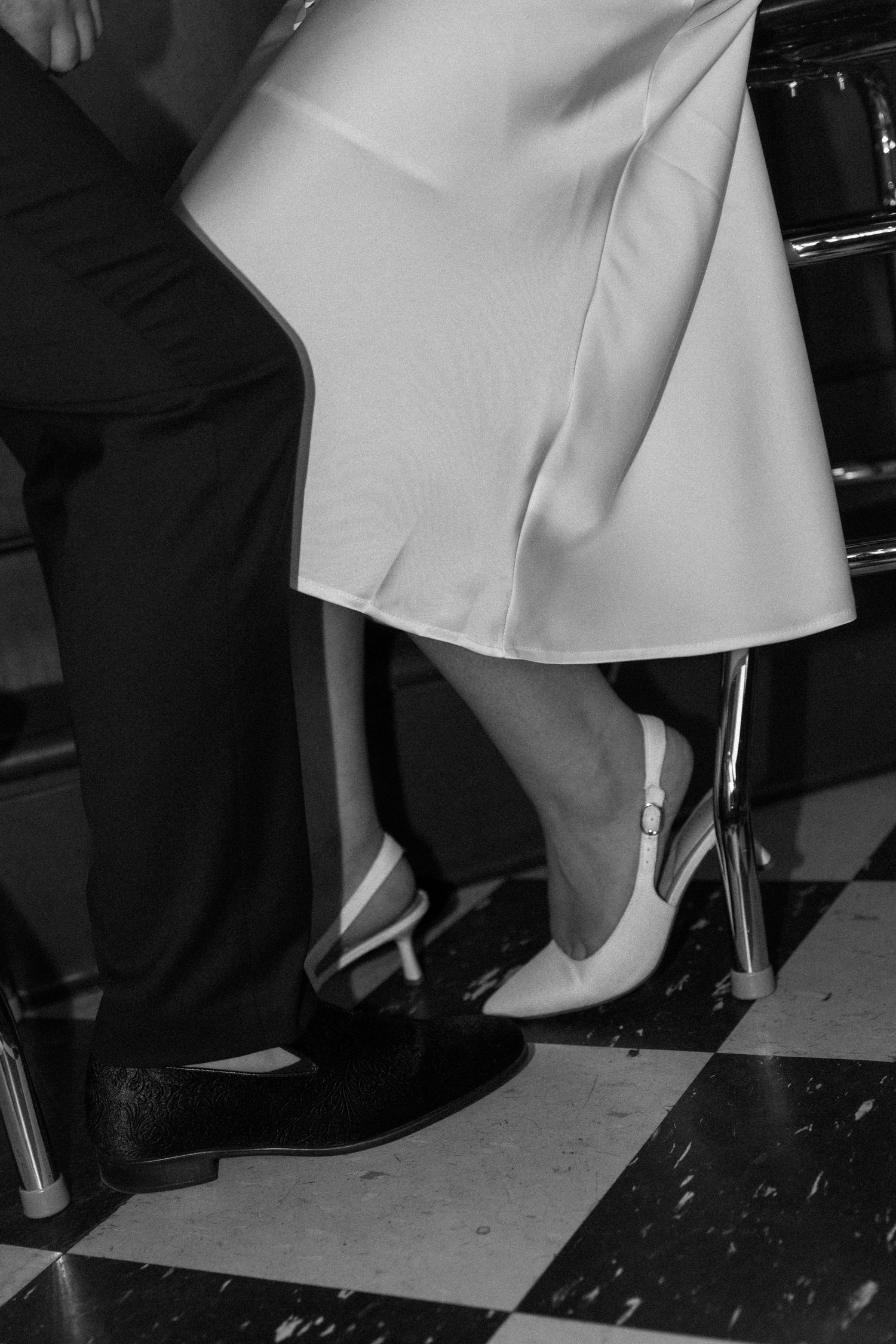 Black and white photo of bride and groom's feet on checkered floor, white heels and dress beside dark suit trousers.