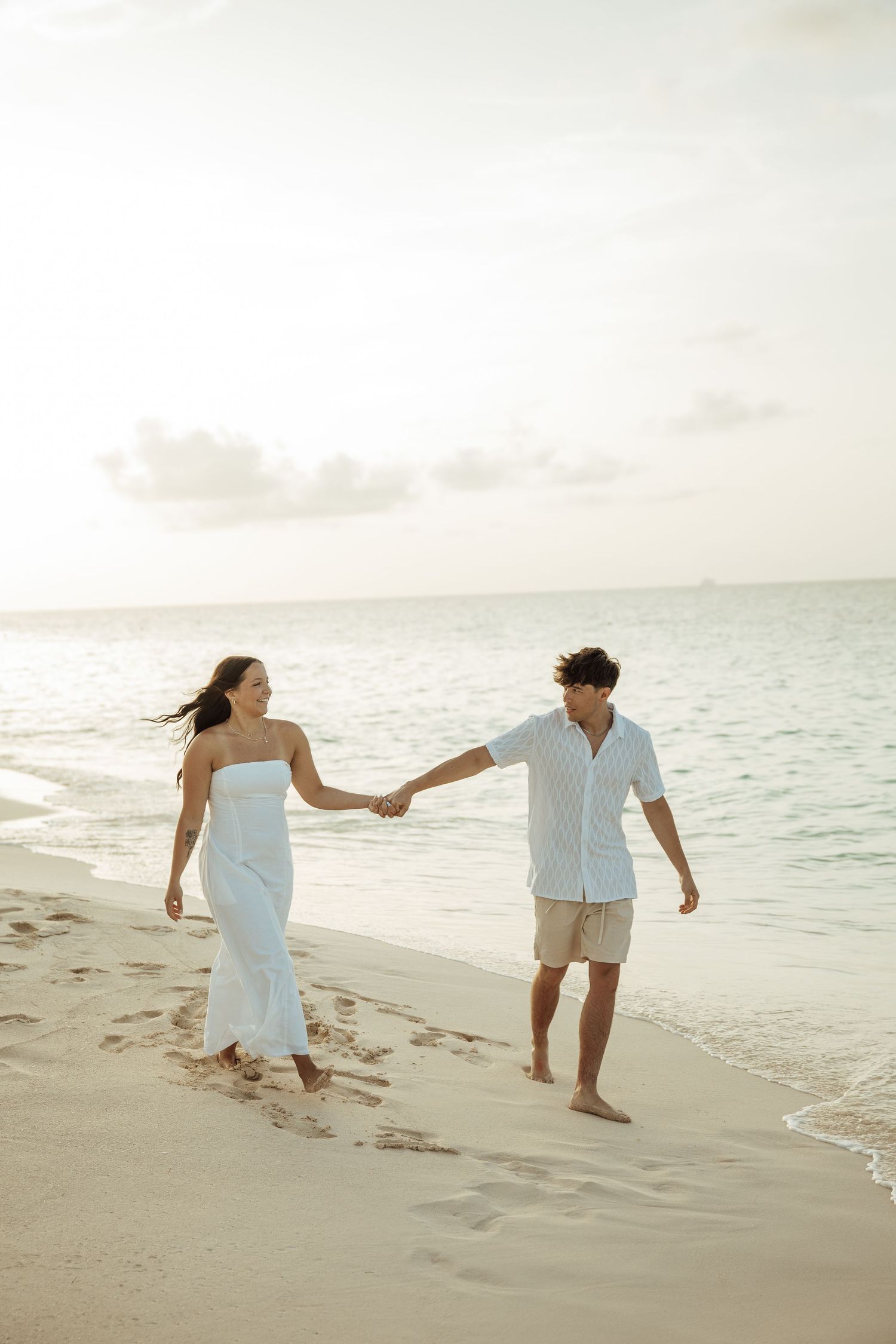 A couple walks hand in hand along a sunlit beach with calm ocean waves in the background.